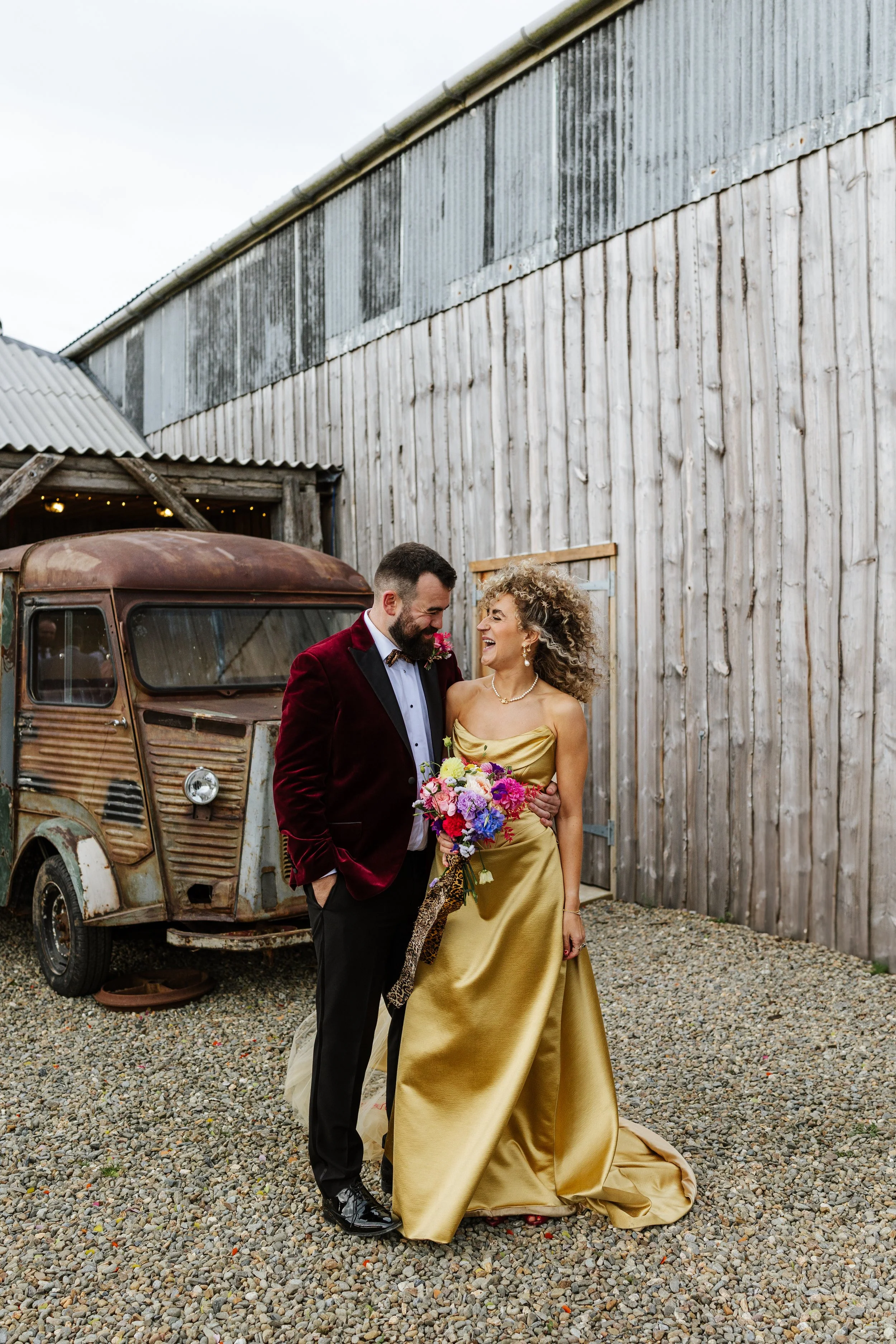 A couple dressed in formal attire standing outside near an old, rusty truck and a weathered wooden barn, smiling and looking at each other.