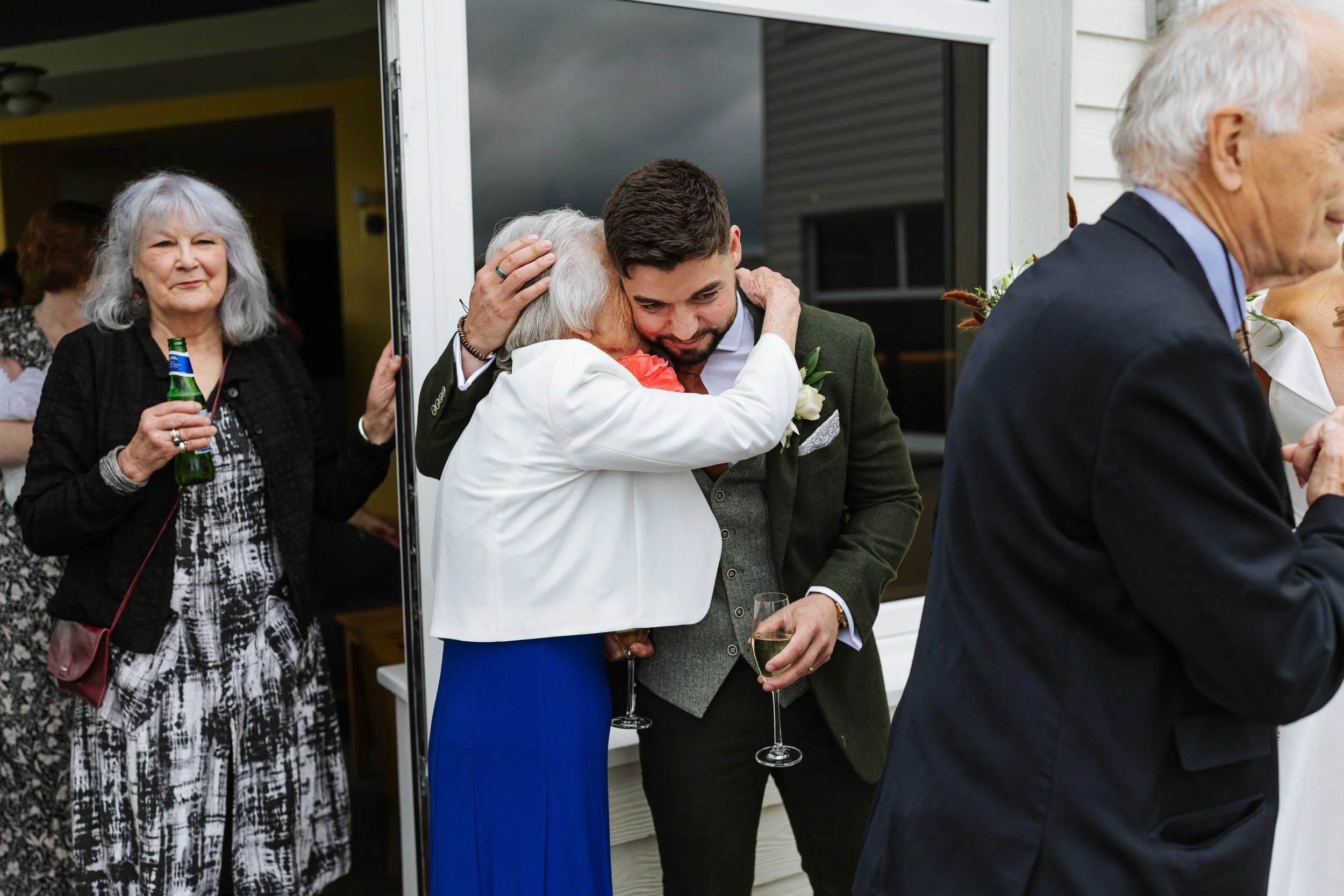 Group of people at a social gathering, with an elderly woman hugging a young man, both holding glasses of wine. Other guests are visible, including a woman holding a beer bottle and a man dressed in a suit. The setting appears to be a porch or patio 