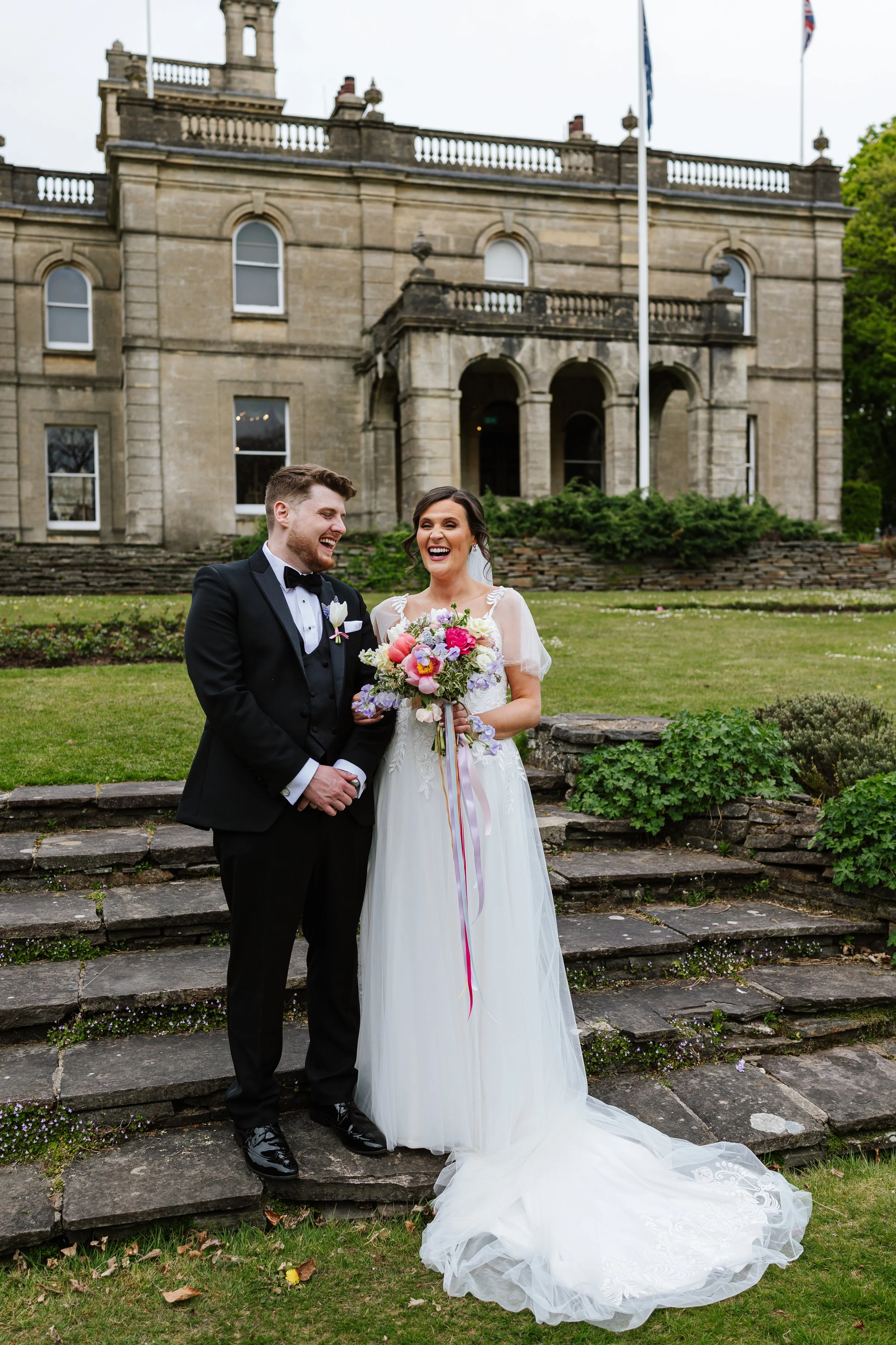 A newlywed couple, a man in a black tuxedo and a woman in a white wedding dress, standing on stone steps in front of a historic stone building with flags. They are smiling and holding a bouquet of flowers, laughing together outdoors.