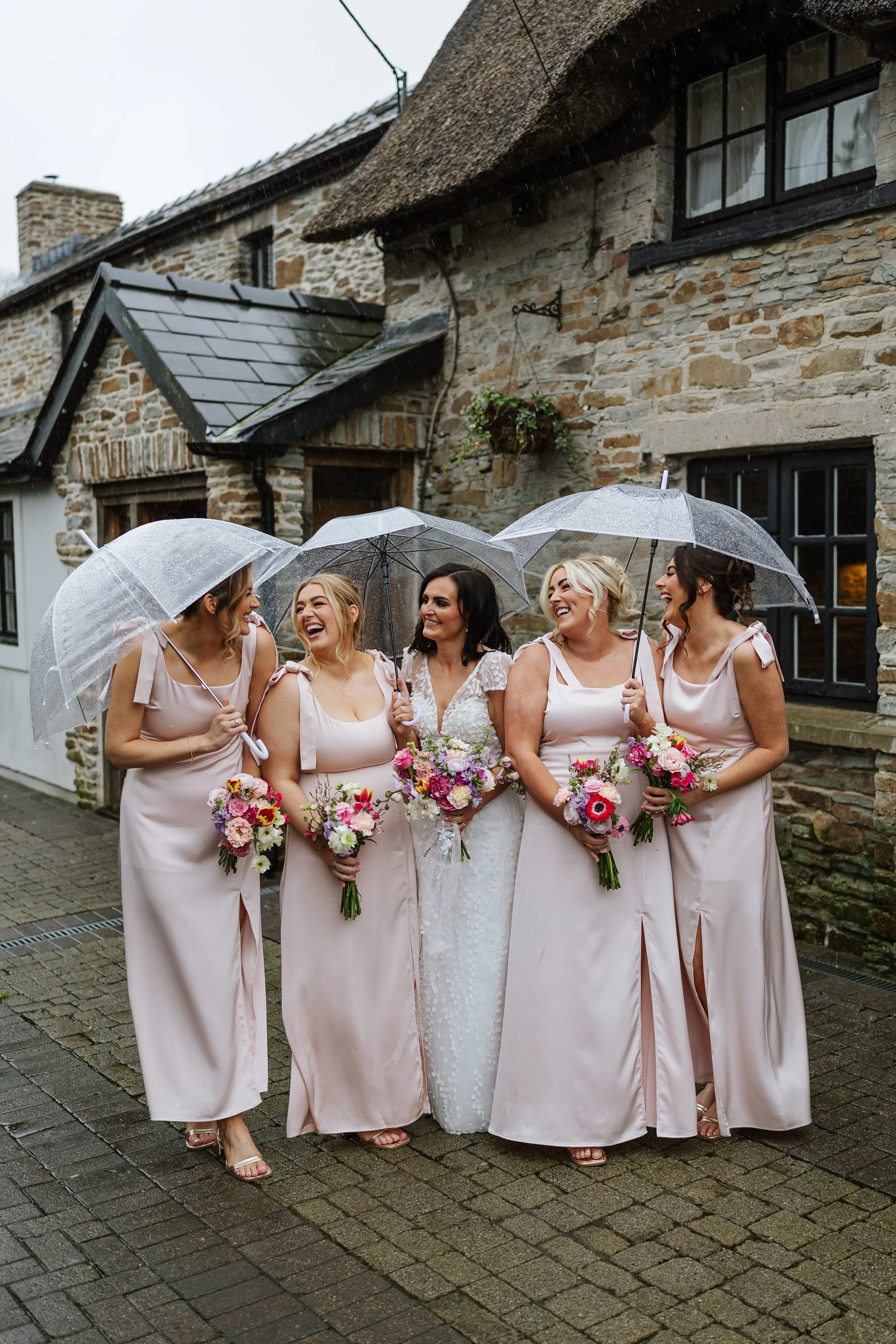 A group of five women, dressed in pink bridesmaid dresses, holding colorful bouquets, smiling, and standing under transparent umbrellas on a rainy day in front of a rustic stone building.