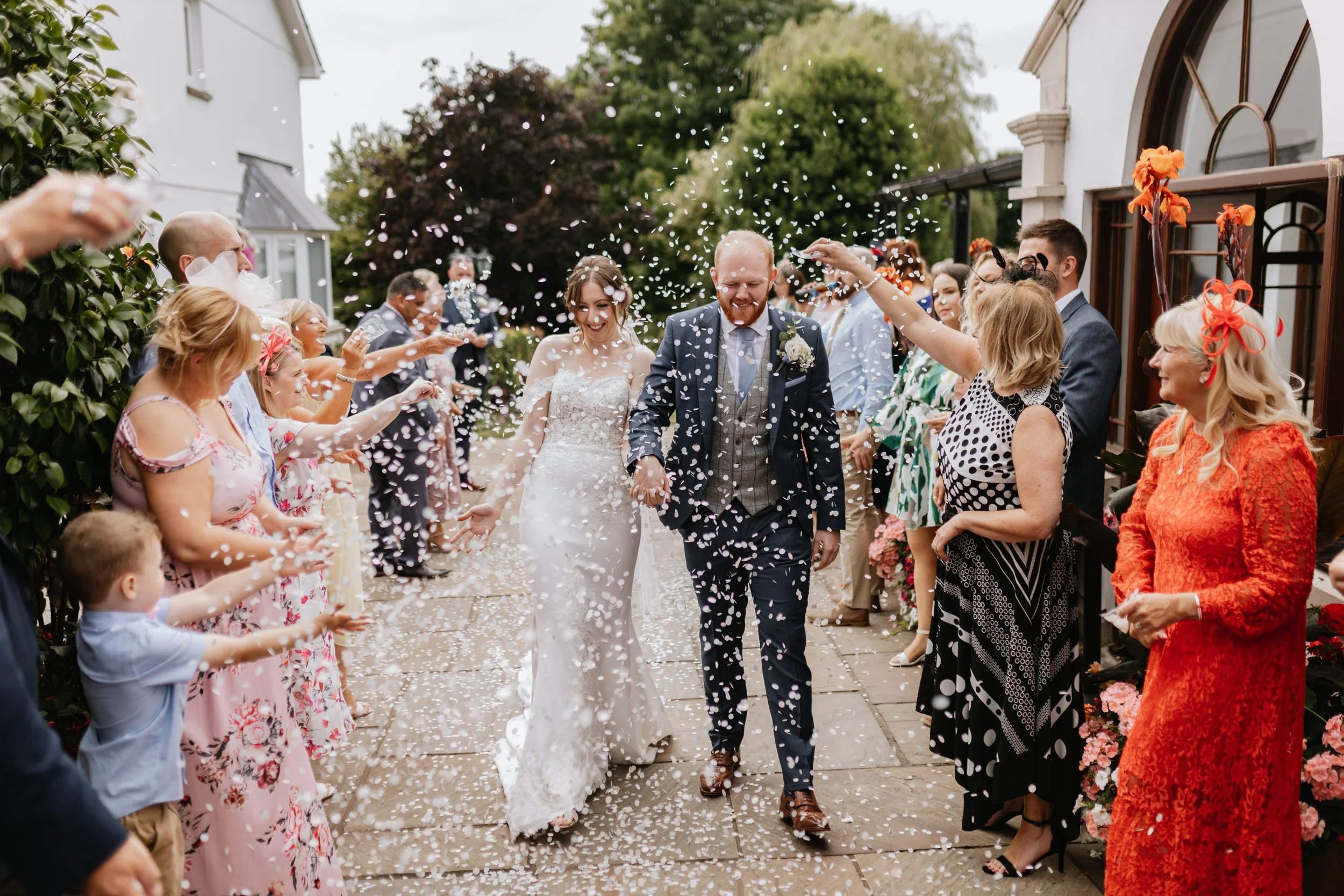 Bride and groom walking through confetti as guests celebrate outside a venue with white walls and brown window frames.
