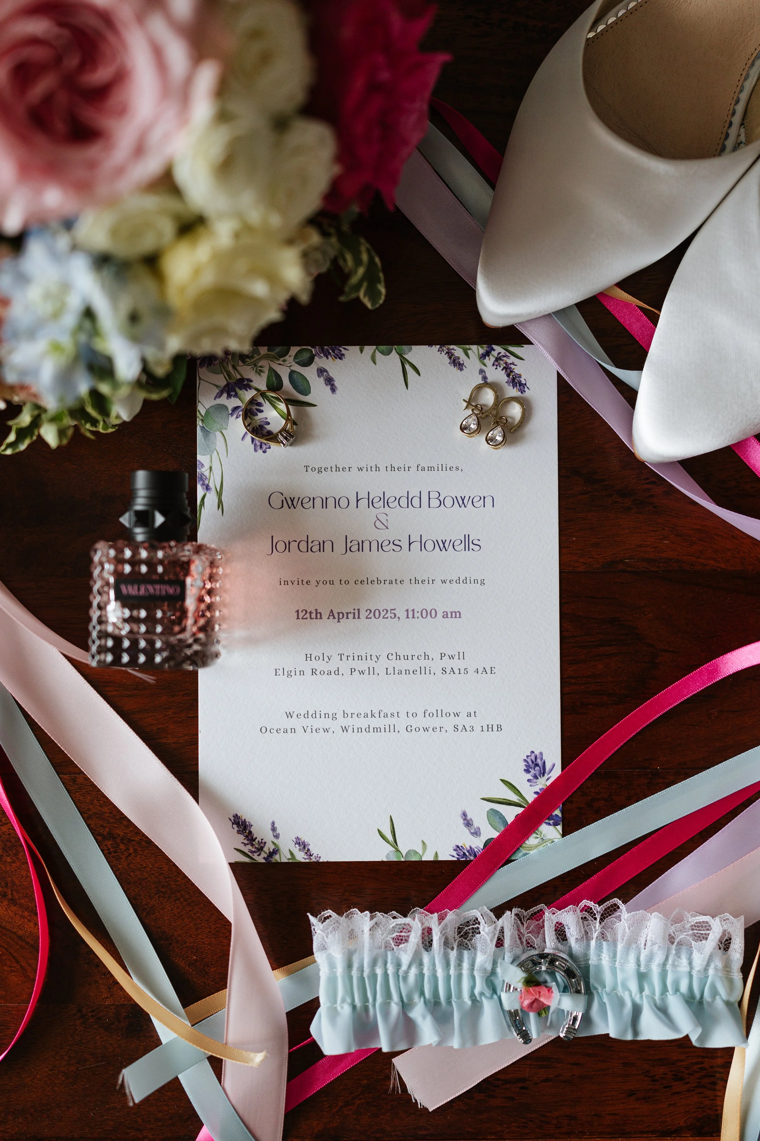 Wedding invitation surrounded by ribbons, flowers, and accessories on a wooden surface.