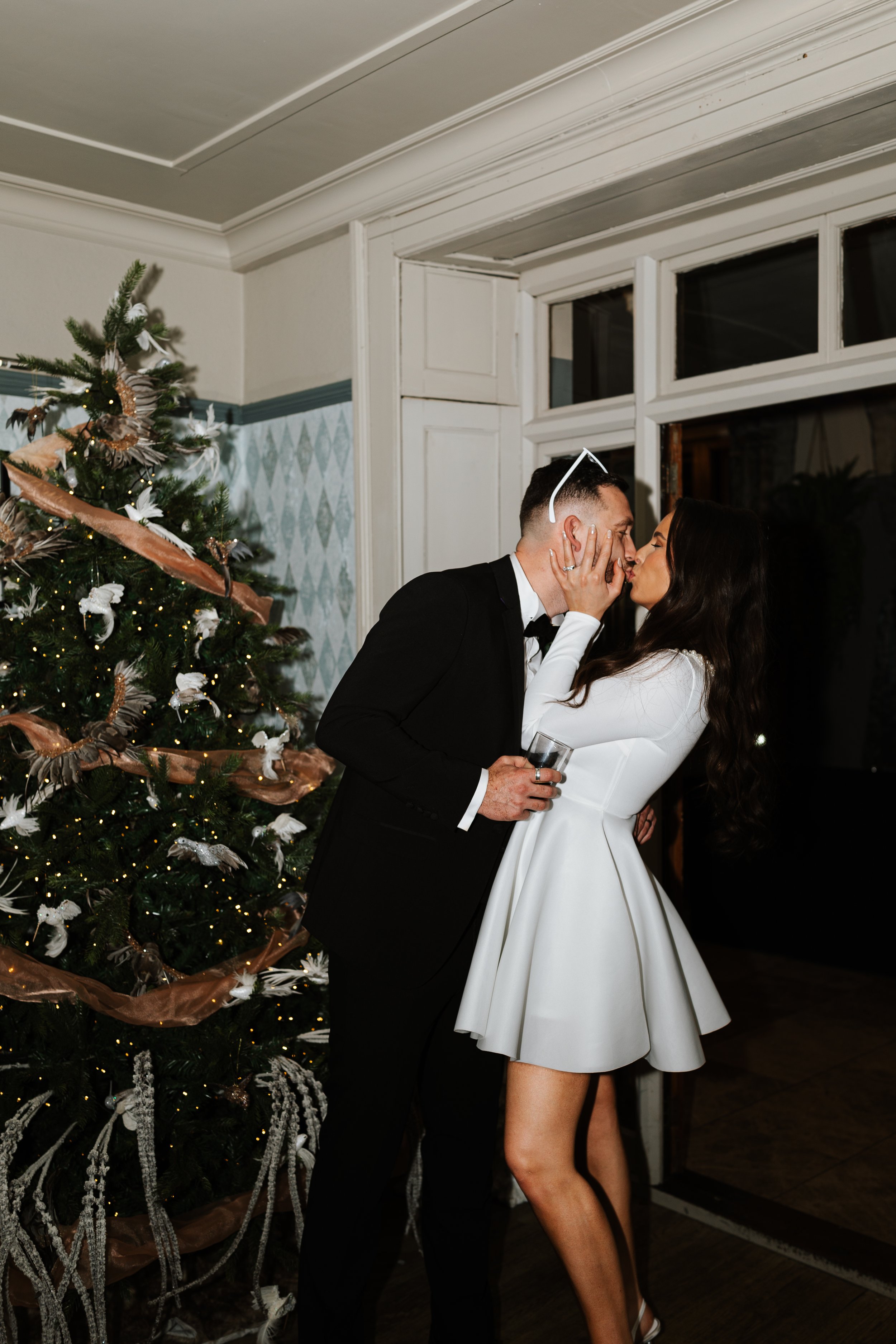 A man and a woman are sharing a kiss at a Christmas party near a decorated Christmas tree, with the woman holding a drink in her hand.