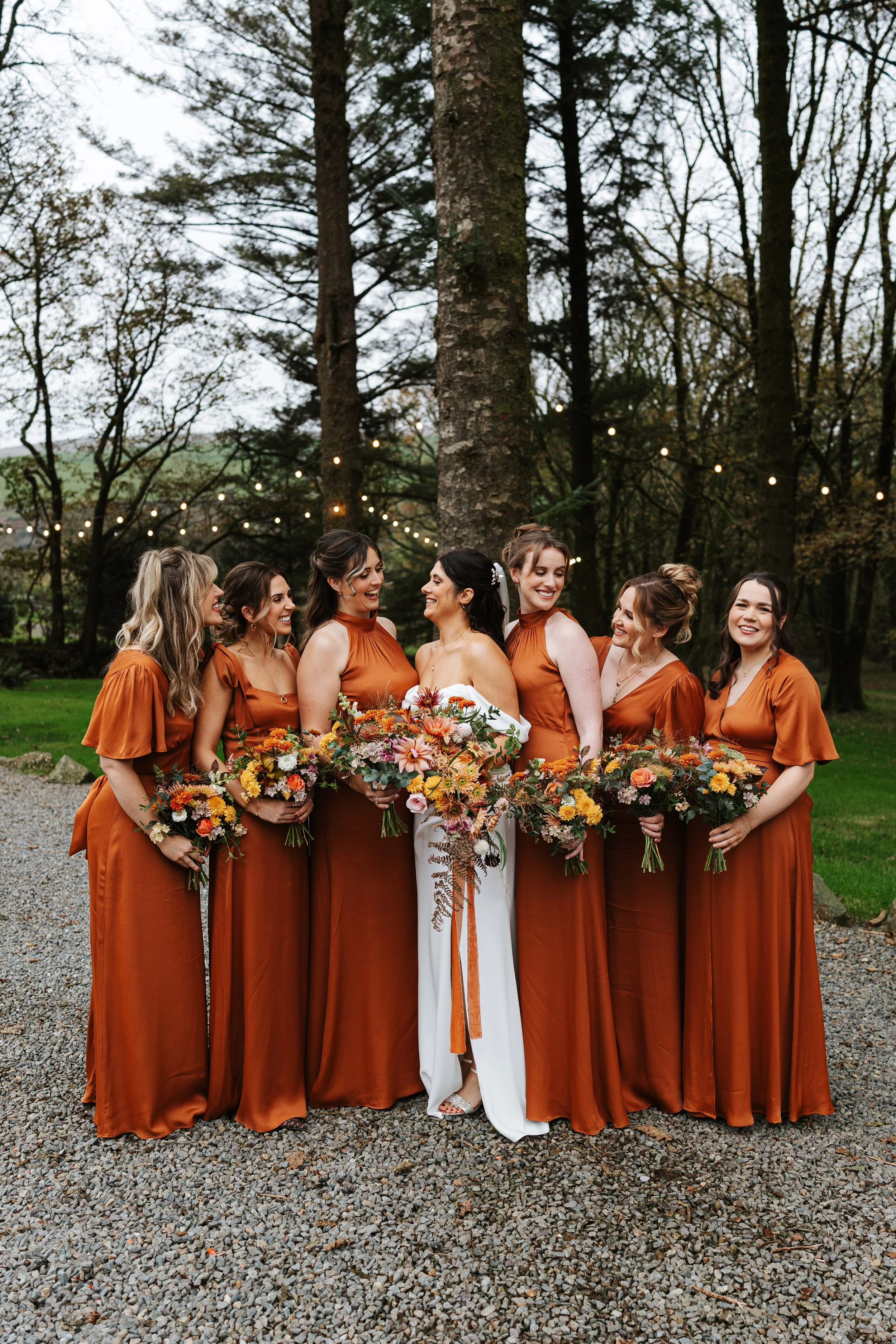 A bride in a white dress surrounded by bridesmaids in rust-colored dresses outdoors, holding bouquets of flowers, with string lights and trees in the background.