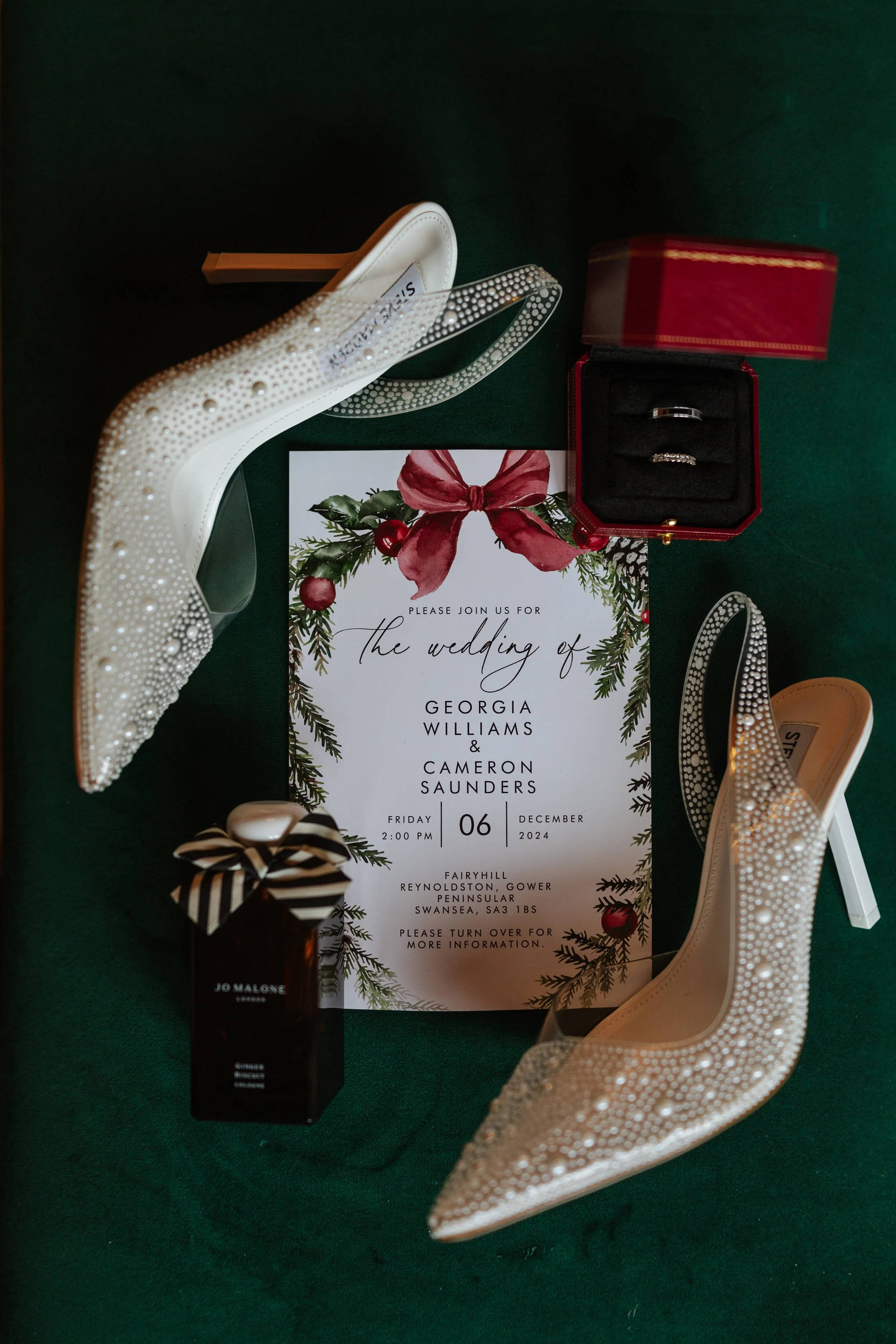 Wedding invitation card with decorative greenery and red bow, a pair of pearl wedding shoes, a jewelry box with rings, a bottle of Jo Malone perfume, and a Christmas greeting card.