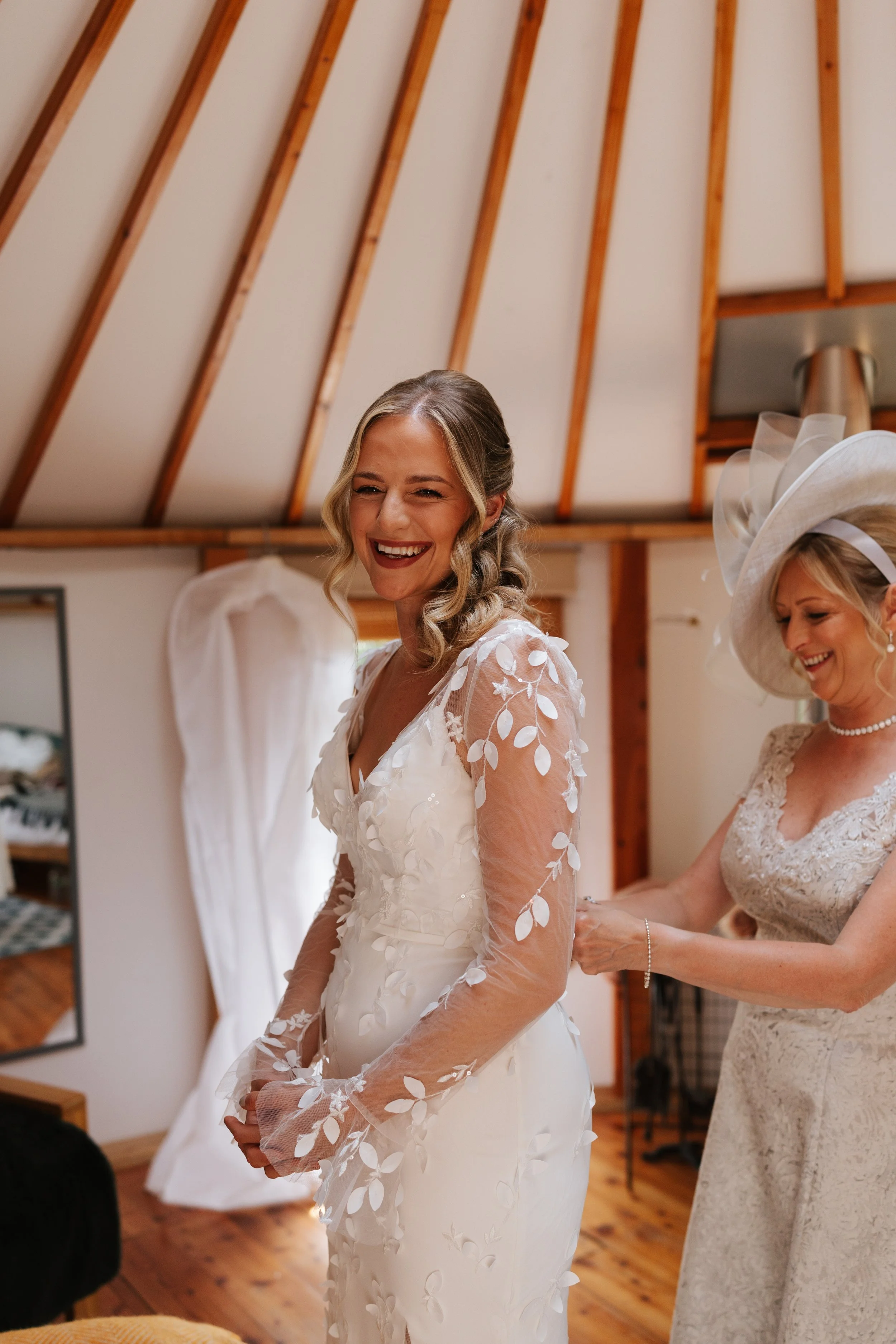 A bride dressed in a white wedding gown with floral lace details is smiling while another woman assists her with her dress in a cozy room with wooden beams.