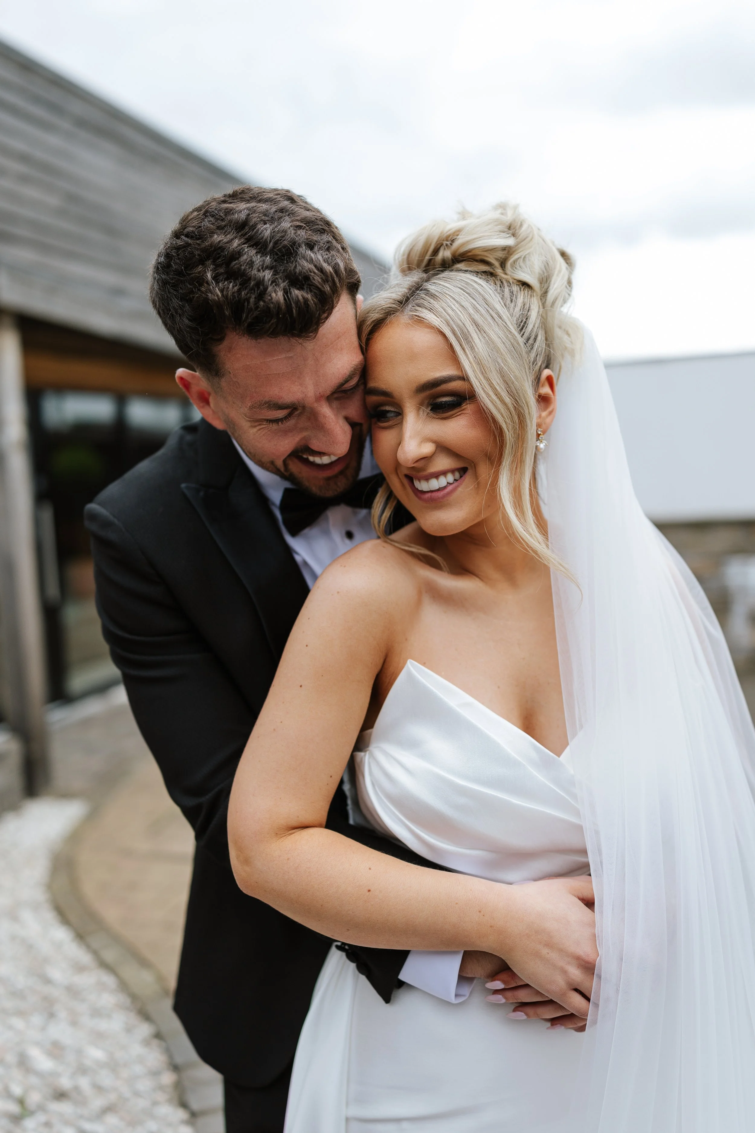 A newlywed couple sharing a joyful moment outdoors, with the groom in a black tuxedo and the bride in a white wedding gown, smiling and holding each other closely.