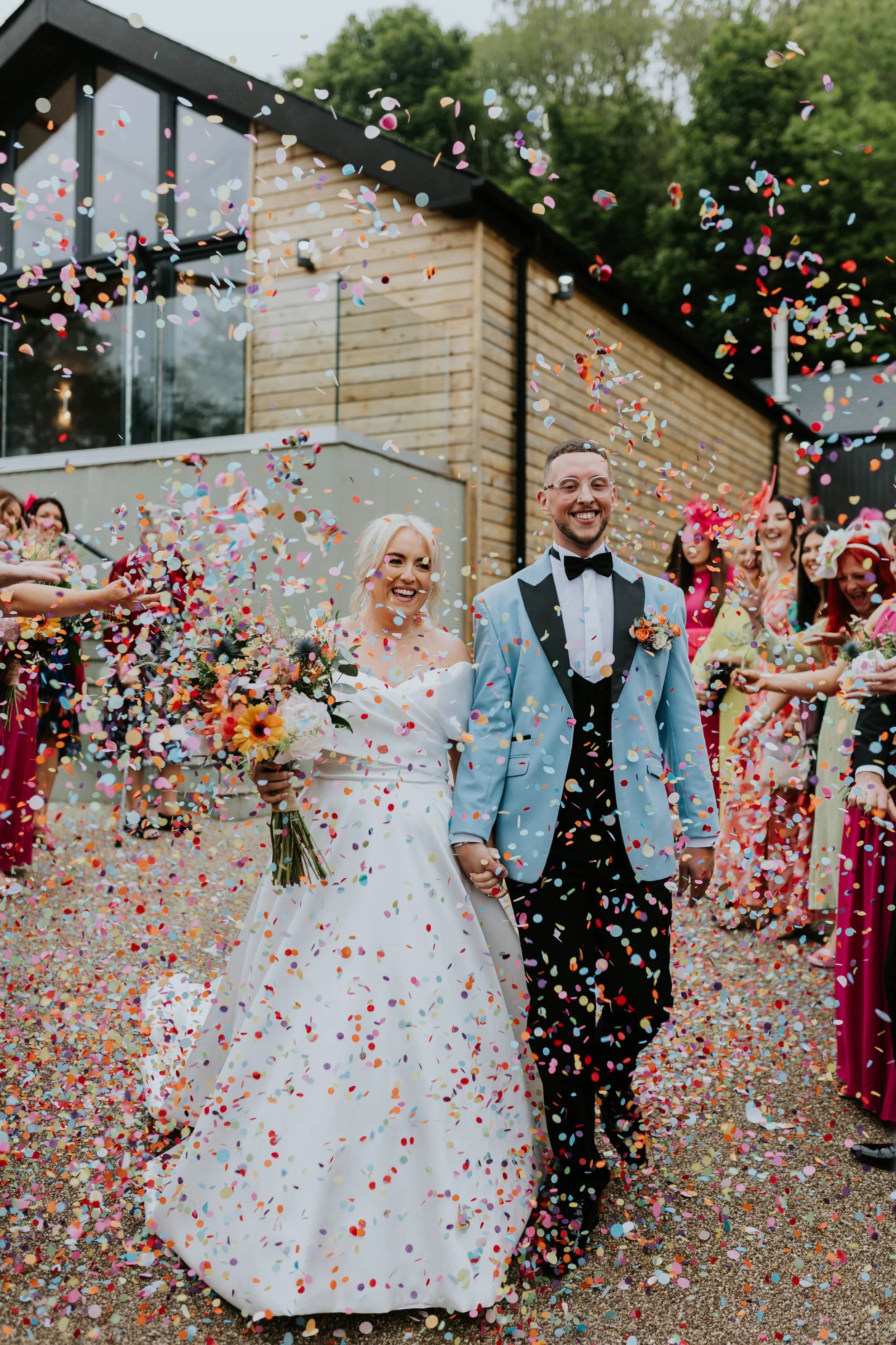 A bride and groom smiling and holding hands as they walk through colorful confetti at their outdoor wedding celebration; the bride is holding a bouquet of flowers, and the groom is wearing a tuxedo with a light blue jacket.