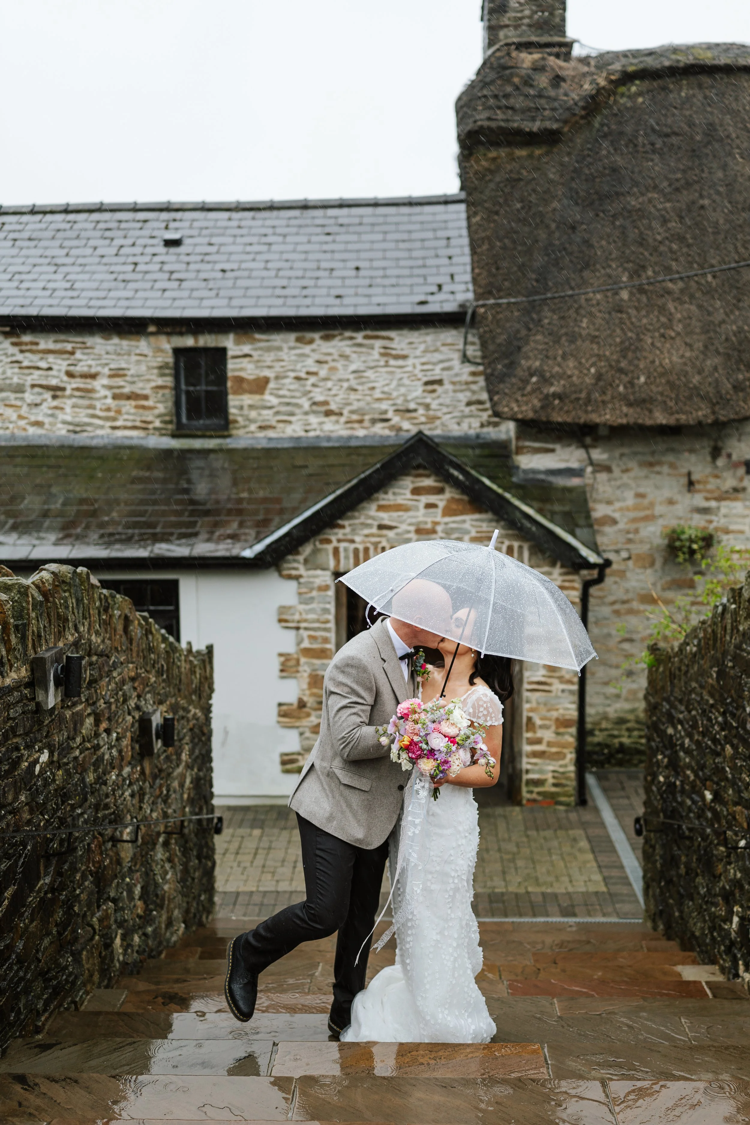A bride and groom share a kiss under a clear umbrella in the rain outside a stone building, with stone pathway and wet stones on the ground.
