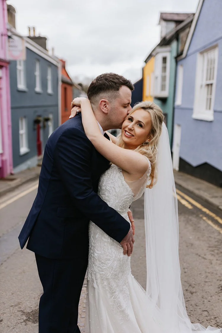 A newlywed couple sharing a loving moment on a colorful street, with the groom kissing the bride on the cheek while she smiles happily and wraps her arms around him.