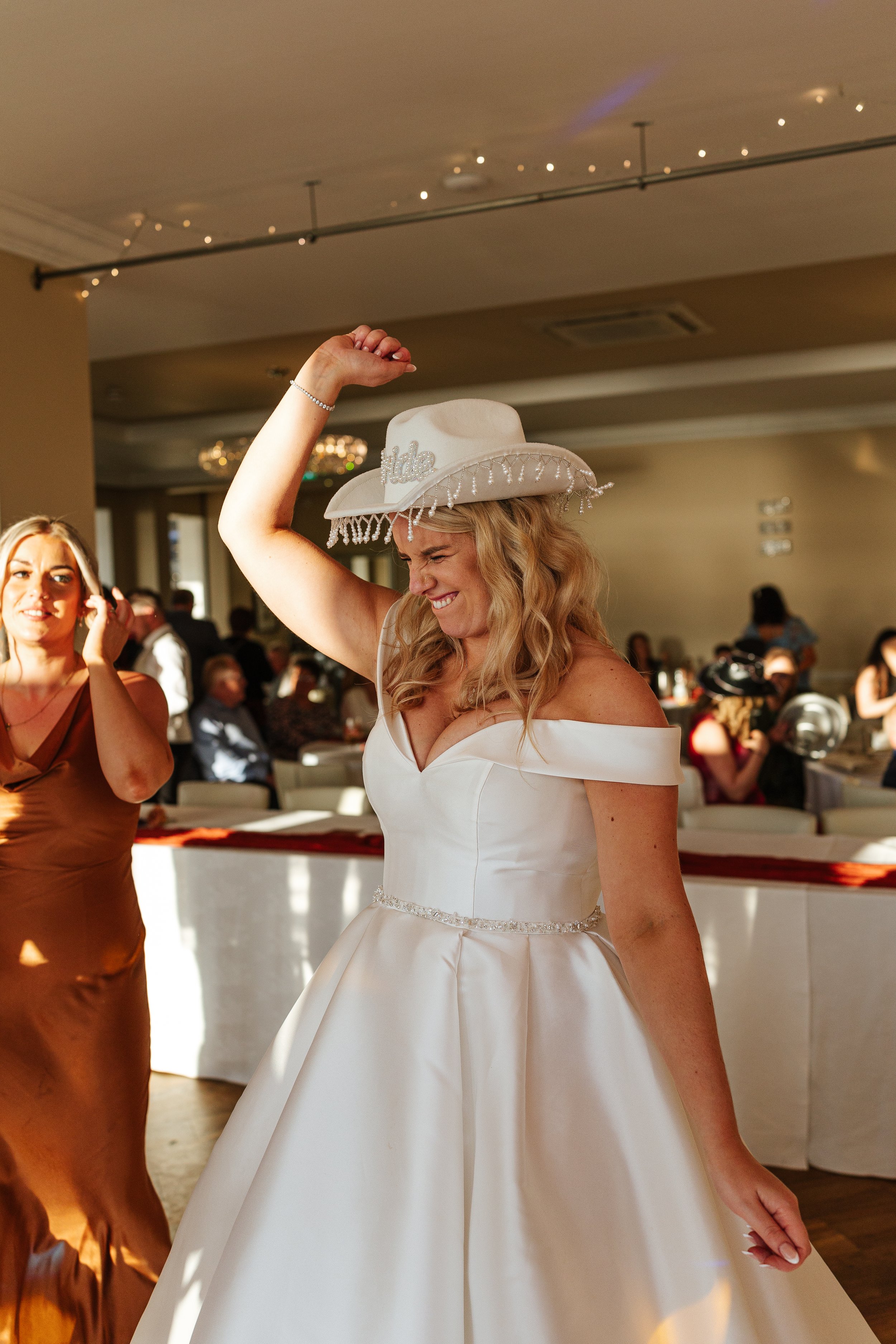 A woman in a white wedding dress and hat dancing at a wedding reception, with guests sitting at tables in the background.