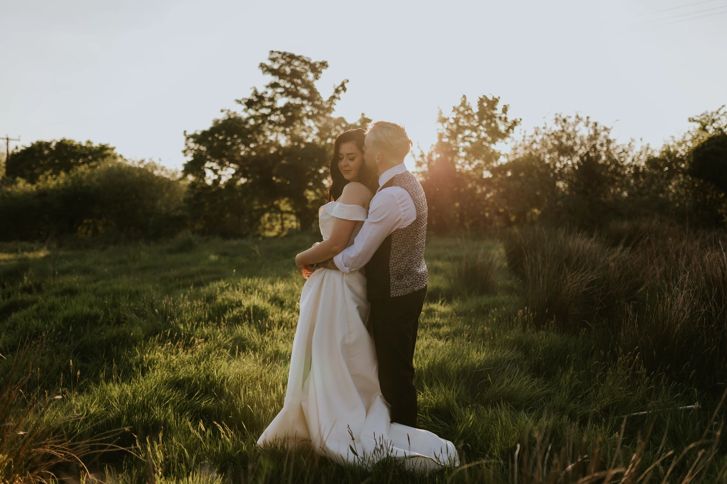 A couple dancing outdoors during sunset, holding each other in a grassy field surrounded by trees.