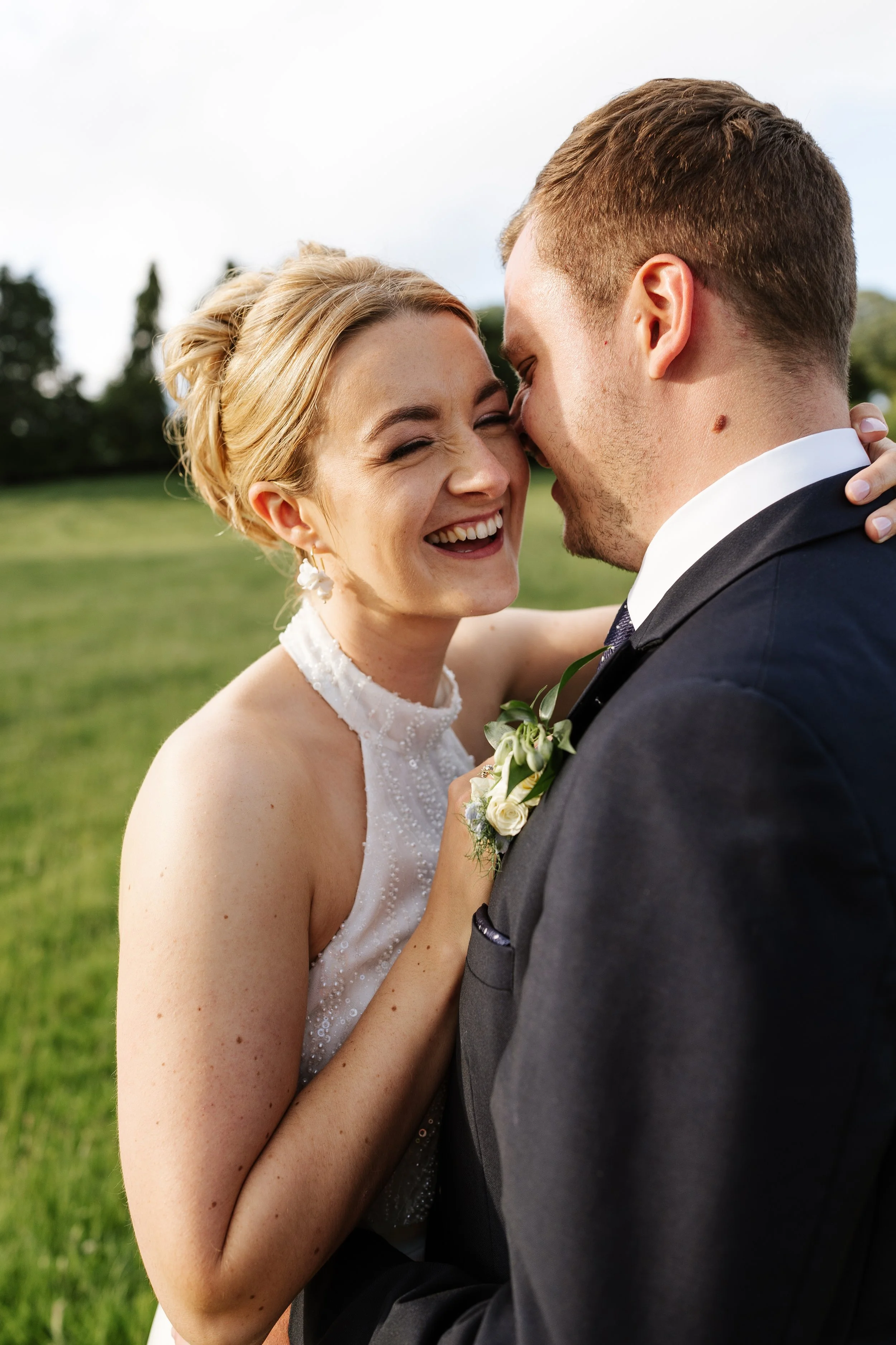A smiling bride and groom embrace outdoors on a sunny day, with green trees and grass in the background.
