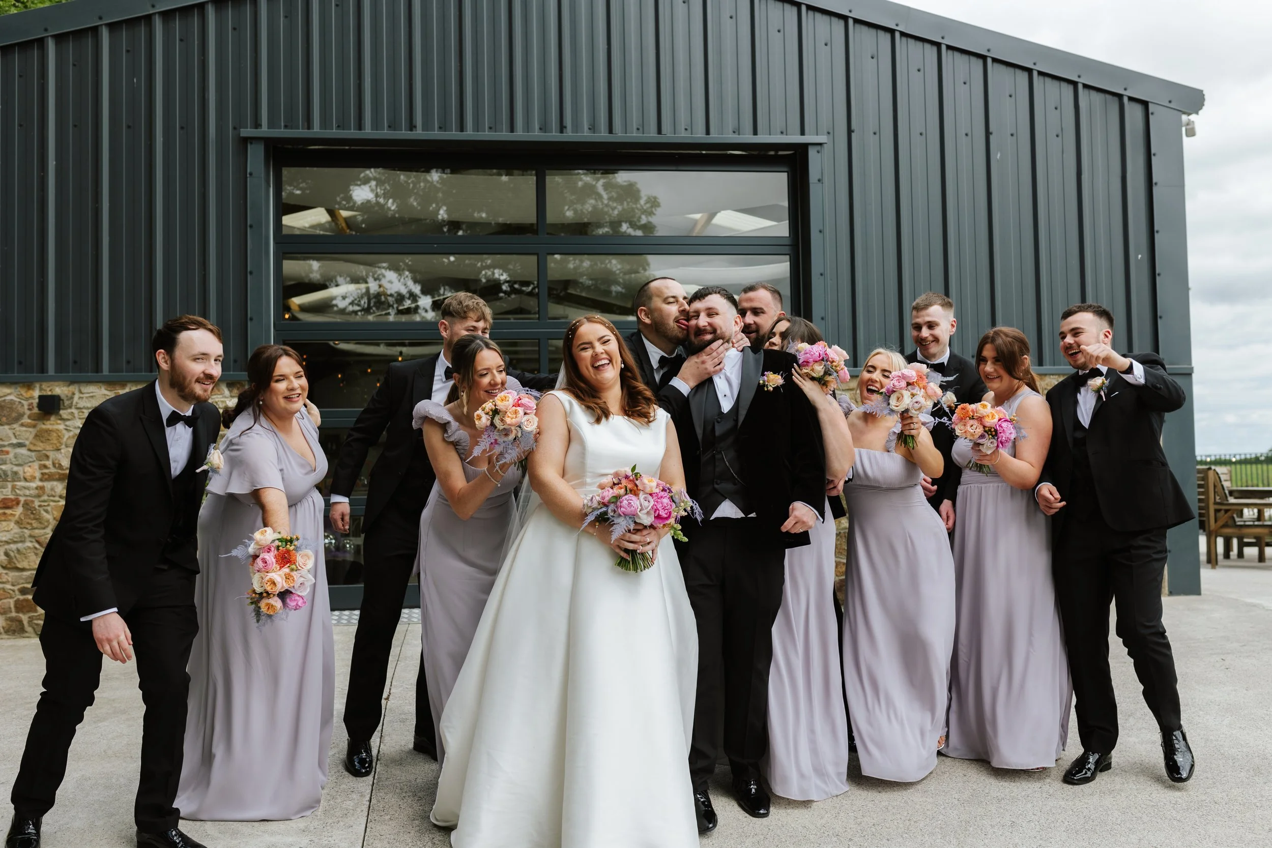 Group of wedding guests, including bride and groom, celebrating outside near a modern building with large windows, during daytime with clouds.