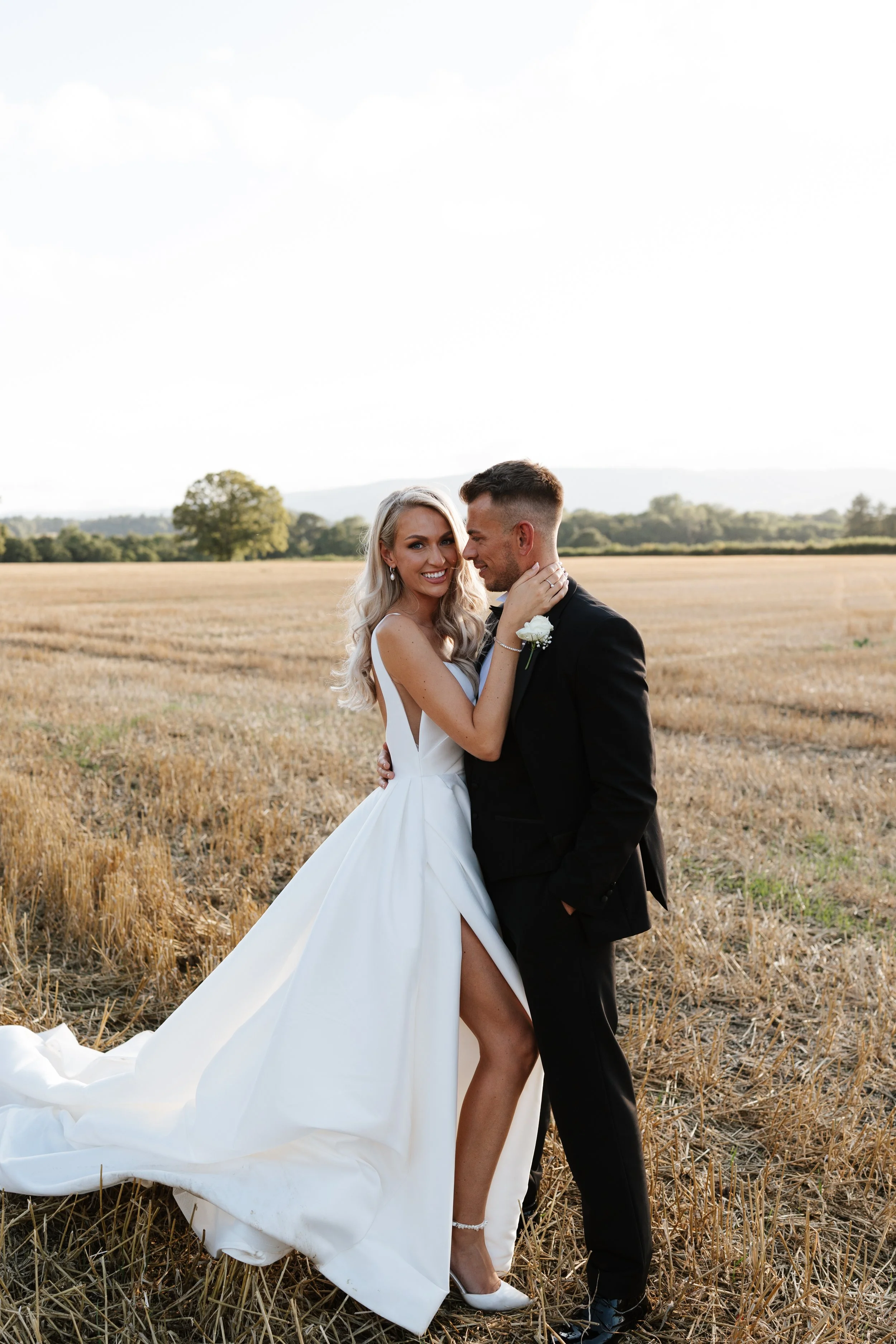 A bride and groom standing in a harvested field, embracing and smiling at each other. The bride is wearing a white gown with a high slit, and the groom is in a black tuxedo. The landscape features distant trees and rolling hills under a partly cloudy