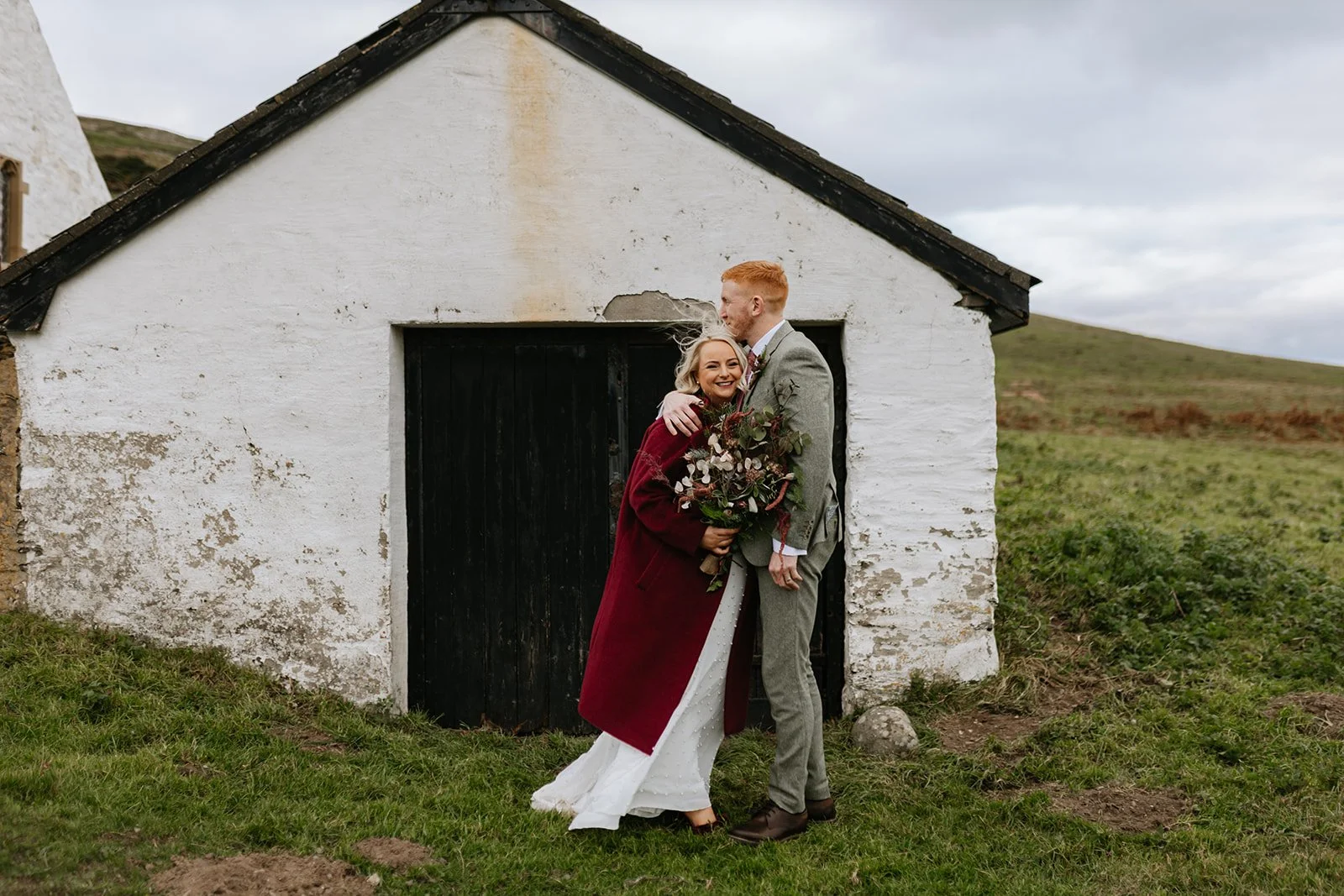 A smiling bride and groom sharing a kiss outside a white rustic building with a black door, holding a bouquet, with grassy fields and hills in the background during overcast weather.