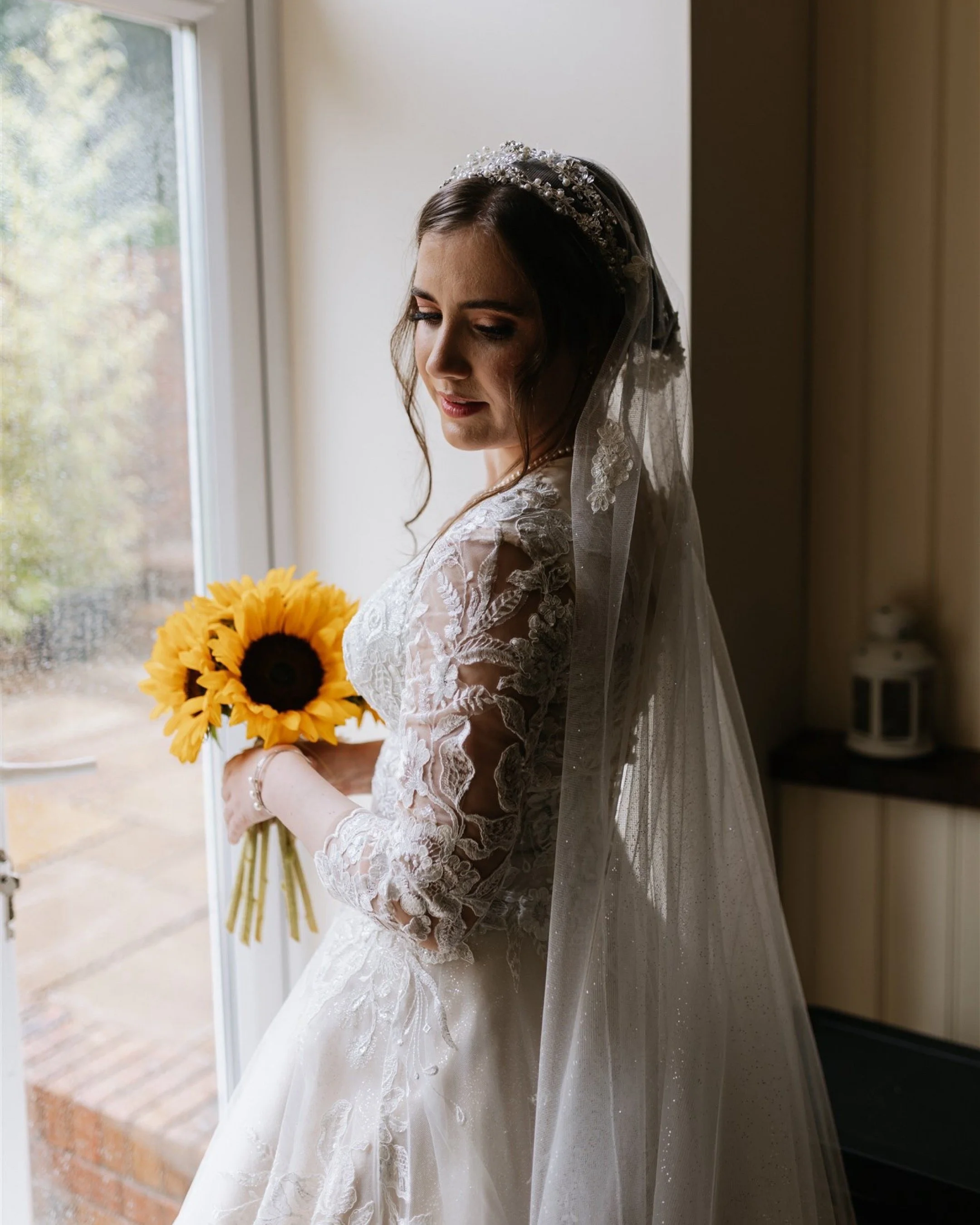 A bride in a white lace wedding dress with a sheer veil and a jeweled headband, holding a bouquet of sunflowers, standing by a window with natural light coming in.