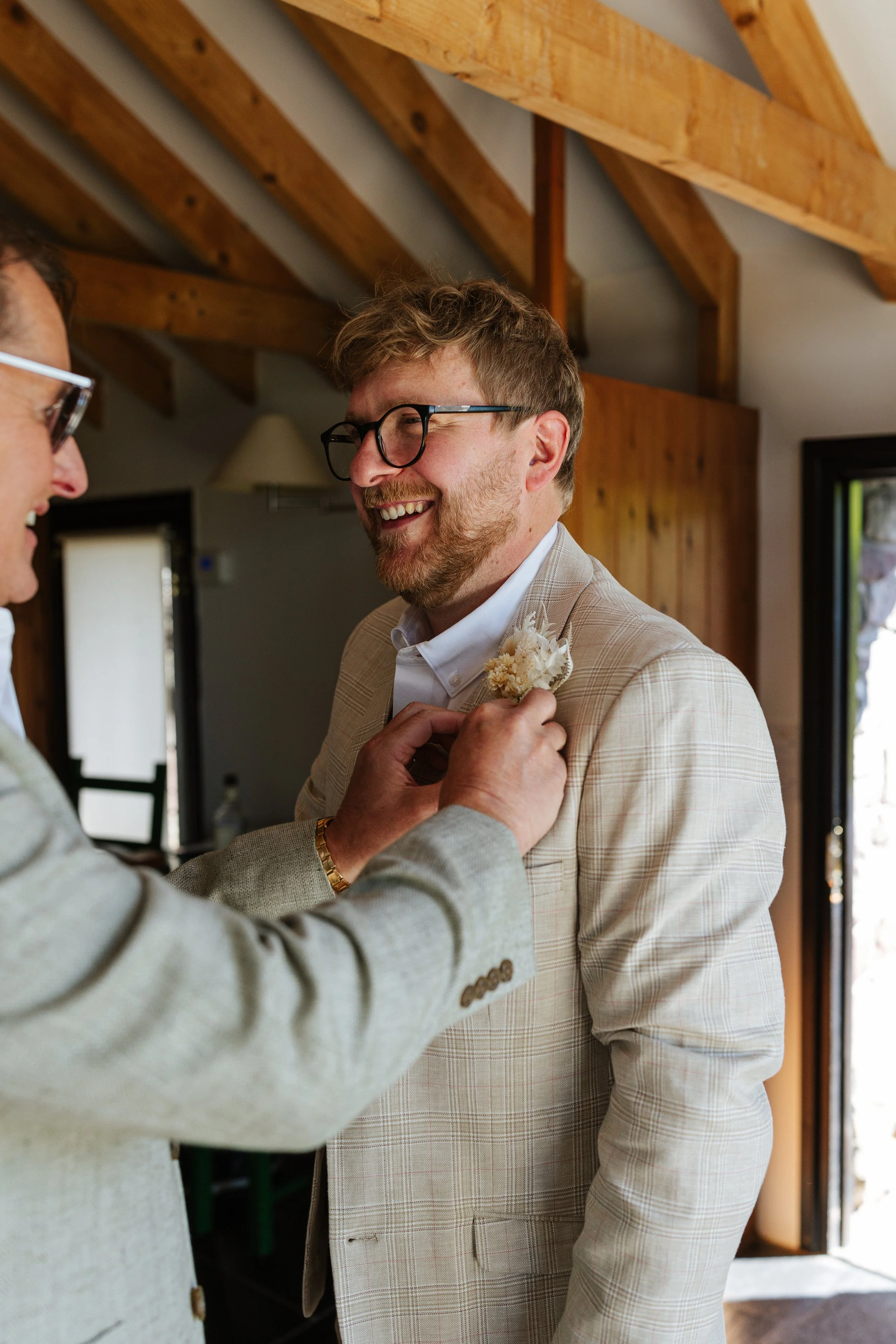 A man in glasses and a light plaid suit, smiling, is being helped by another person with a light-colored blazer to put on a boutonniere.