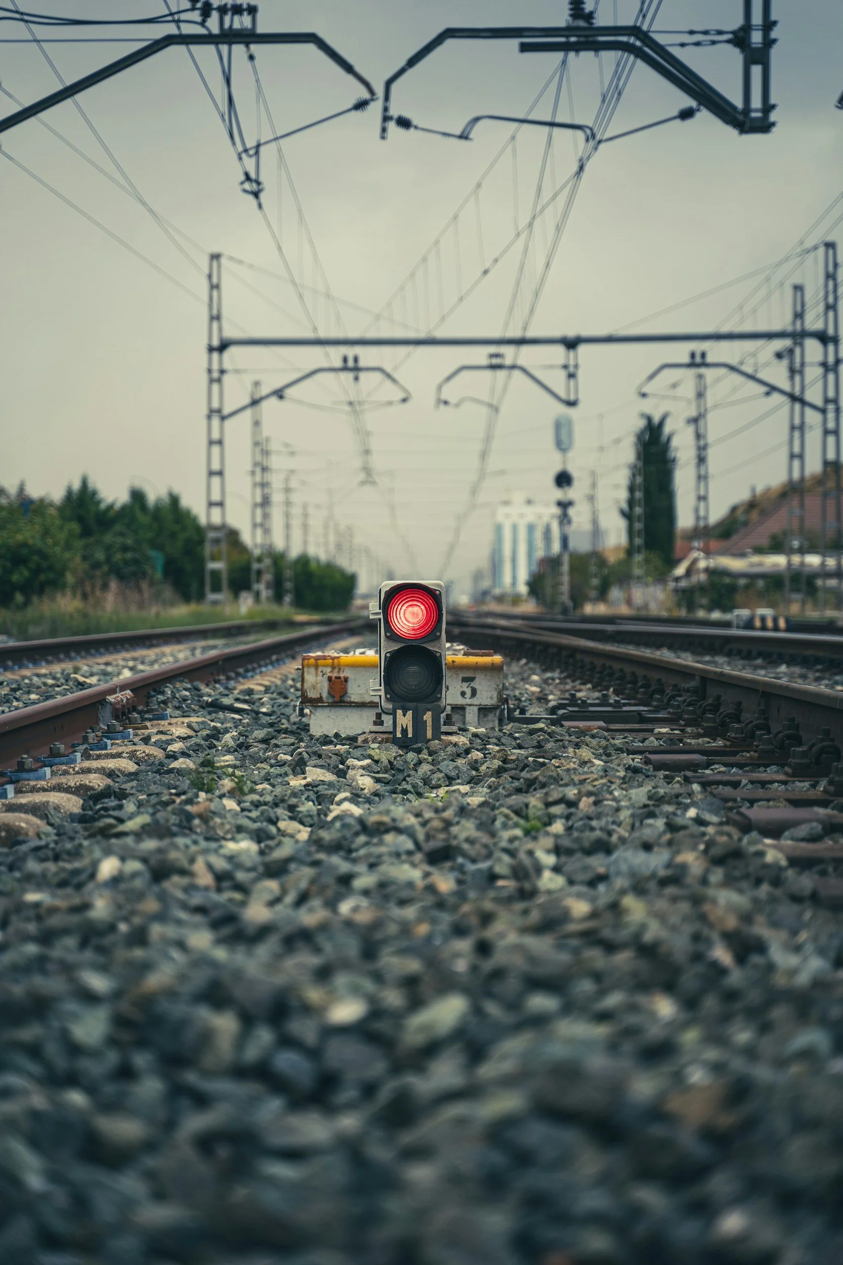 A railway track with a red signal indicating stop, surrounded by gravel and overhead electric wires, with trees and buildings in the distance.