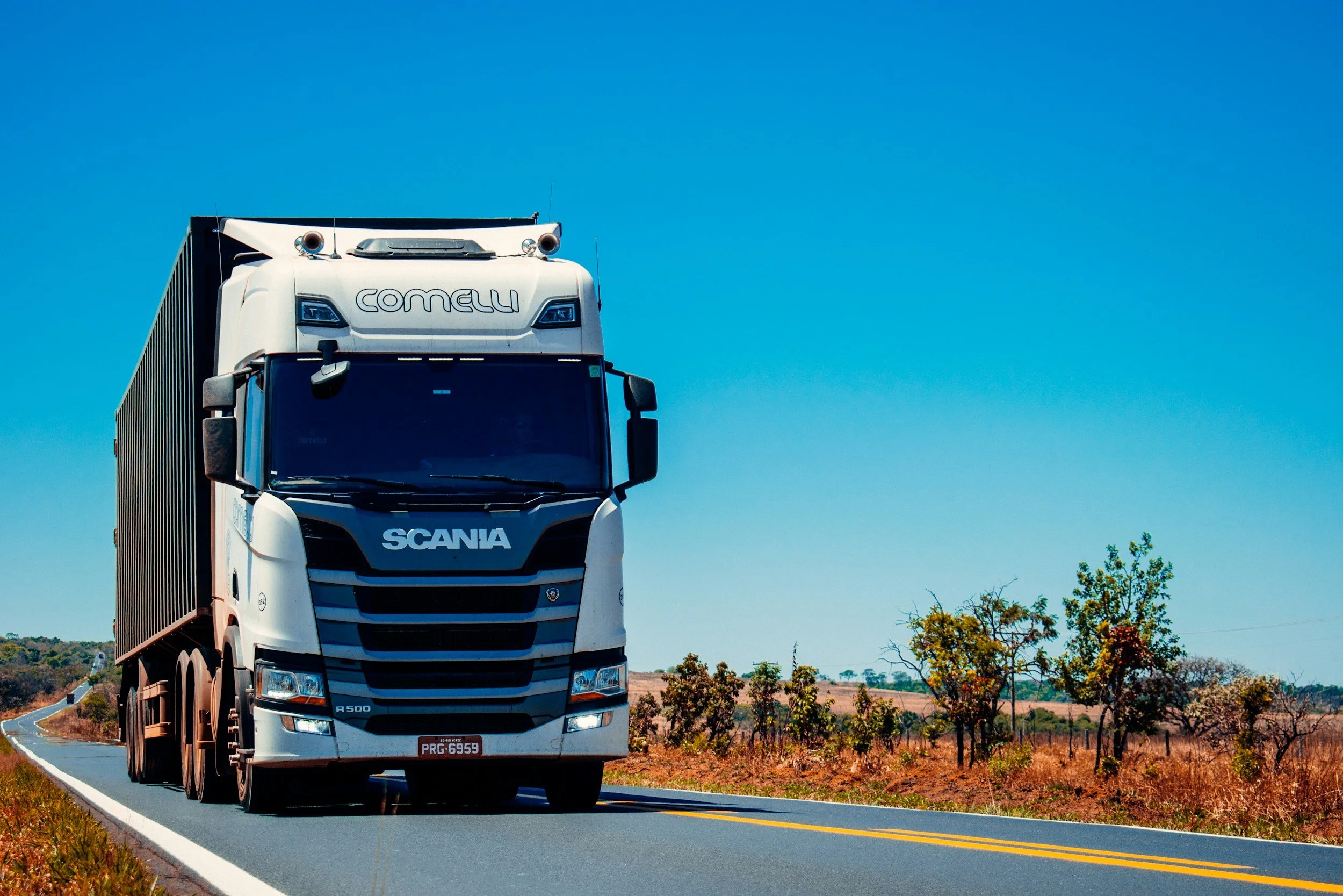 A large white Scania semi-truck driving on a two-lane rural road with a clear blue sky and sparse trees on dry land.