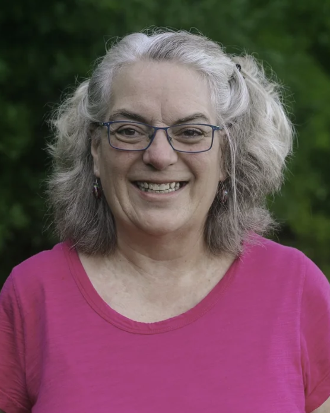 A smiling man in a pink checkered suit, white shirt, and pink tie stands in front of a dark, cityscape background.
