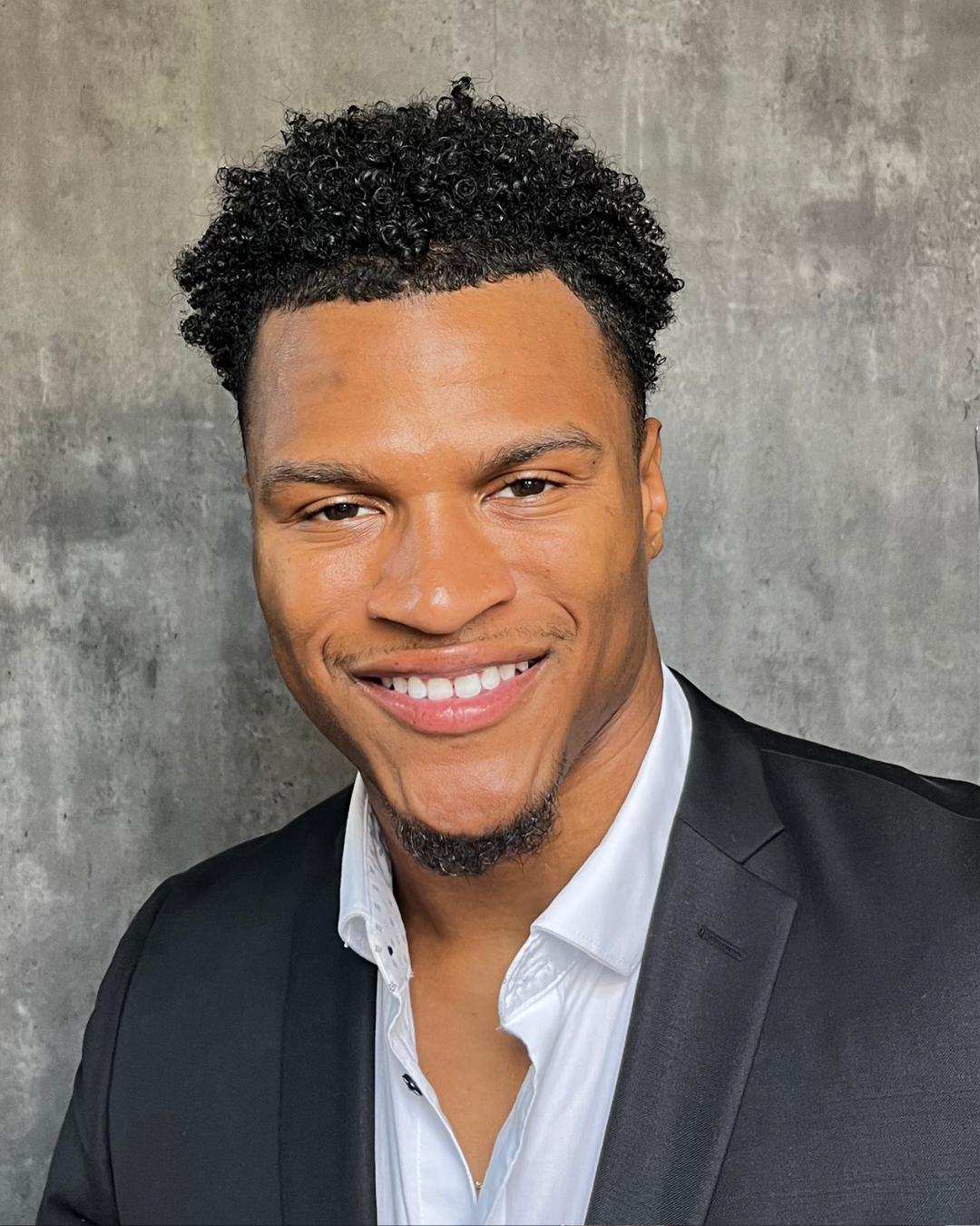 A smiling man in a pink checkered suit, white shirt, and pink tie stands in front of a dark, cityscape background.