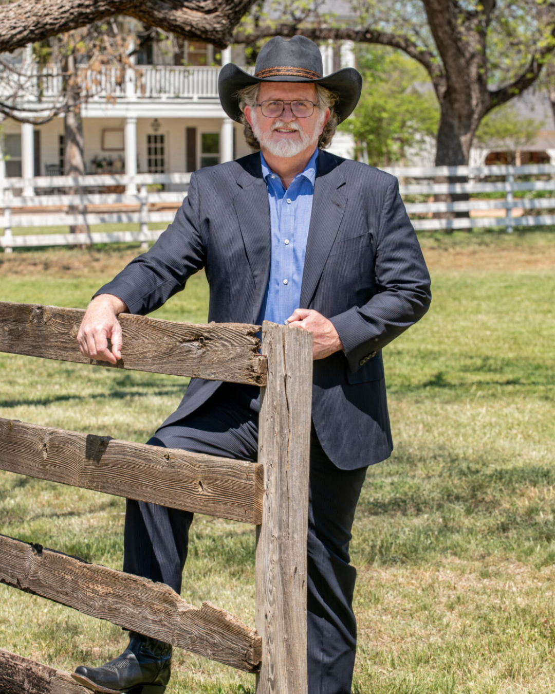 A smiling man in a pink checkered suit, white shirt, and pink tie stands in front of a dark, cityscape background.