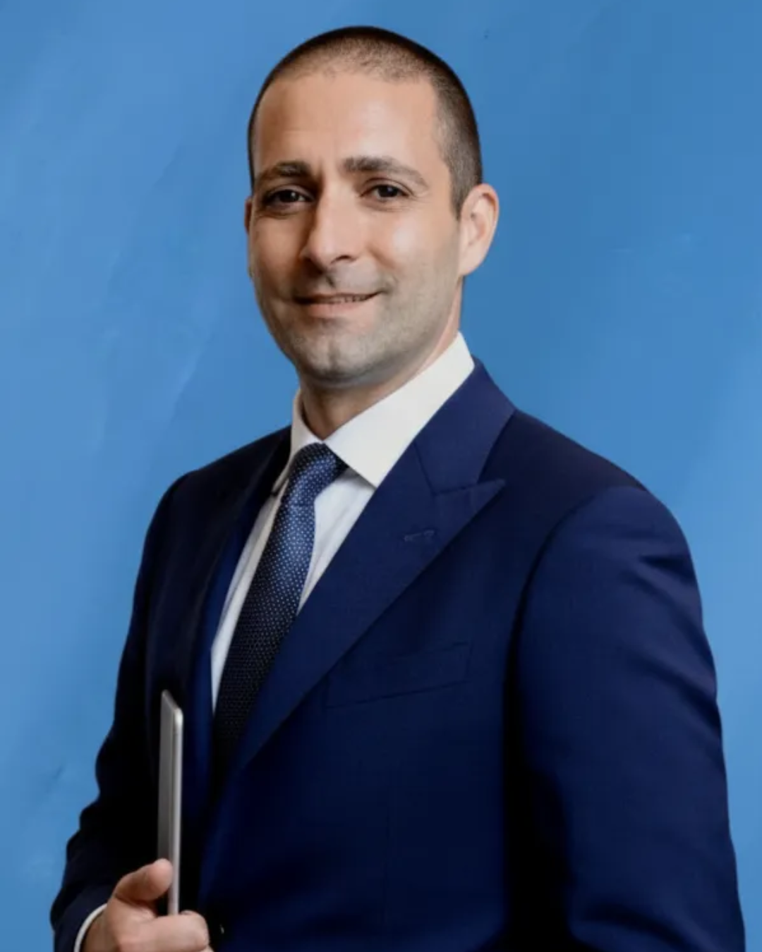 A smiling man in a pink checkered suit, white shirt, and pink tie stands in front of a dark, cityscape background.