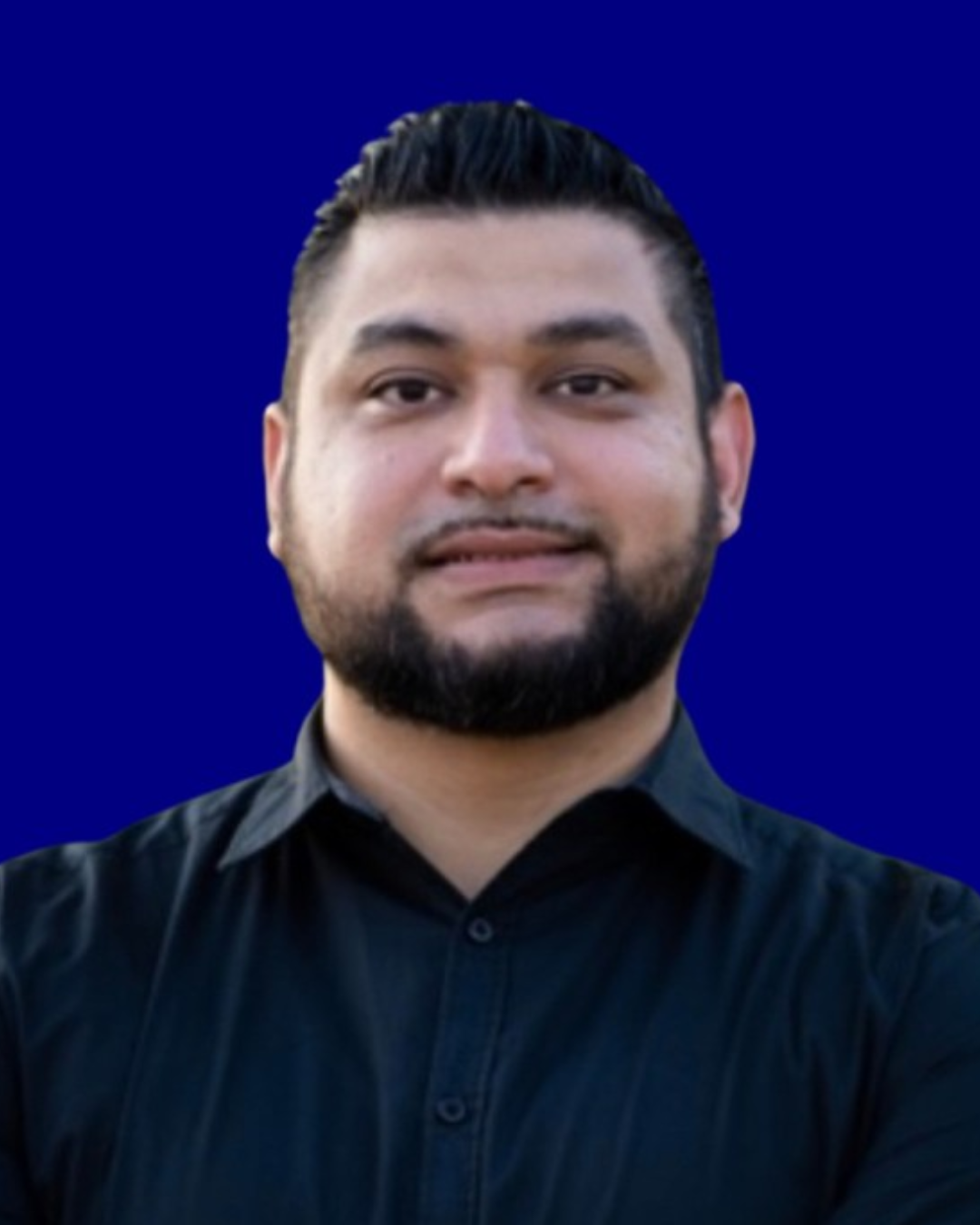 A smiling man in a pink checkered suit, white shirt, and pink tie stands in front of a dark, cityscape background.