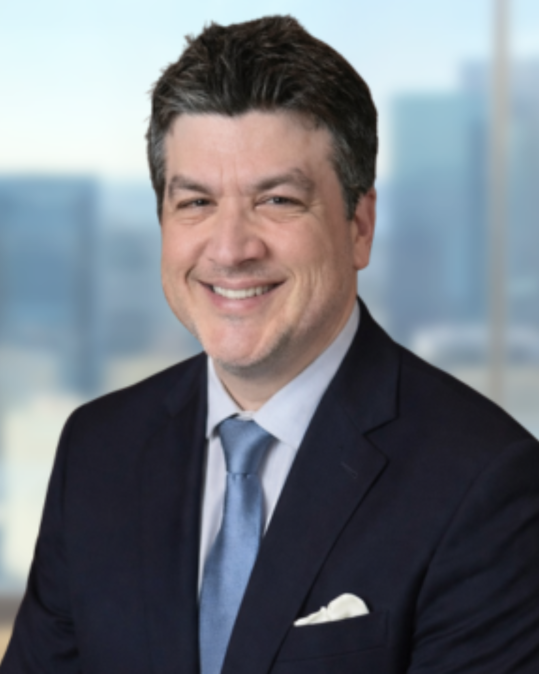 A smiling man in a pink checkered suit, white shirt, and pink tie stands in front of a dark, cityscape background.