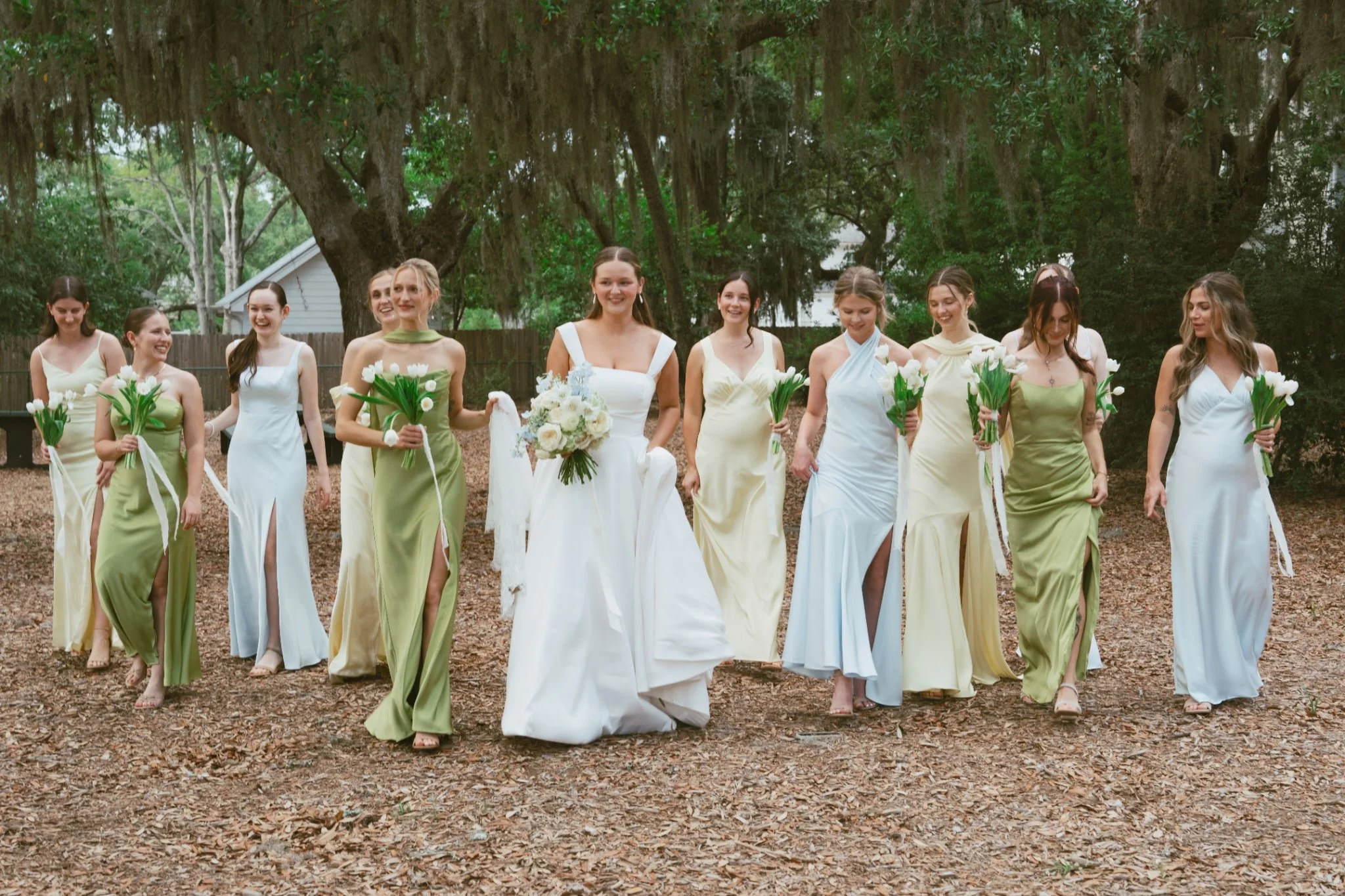 A group of women walking outdoors in a wooded area, dressed in light-colored dresses, with the bride in a white dress holding a bouquet of flowers.
