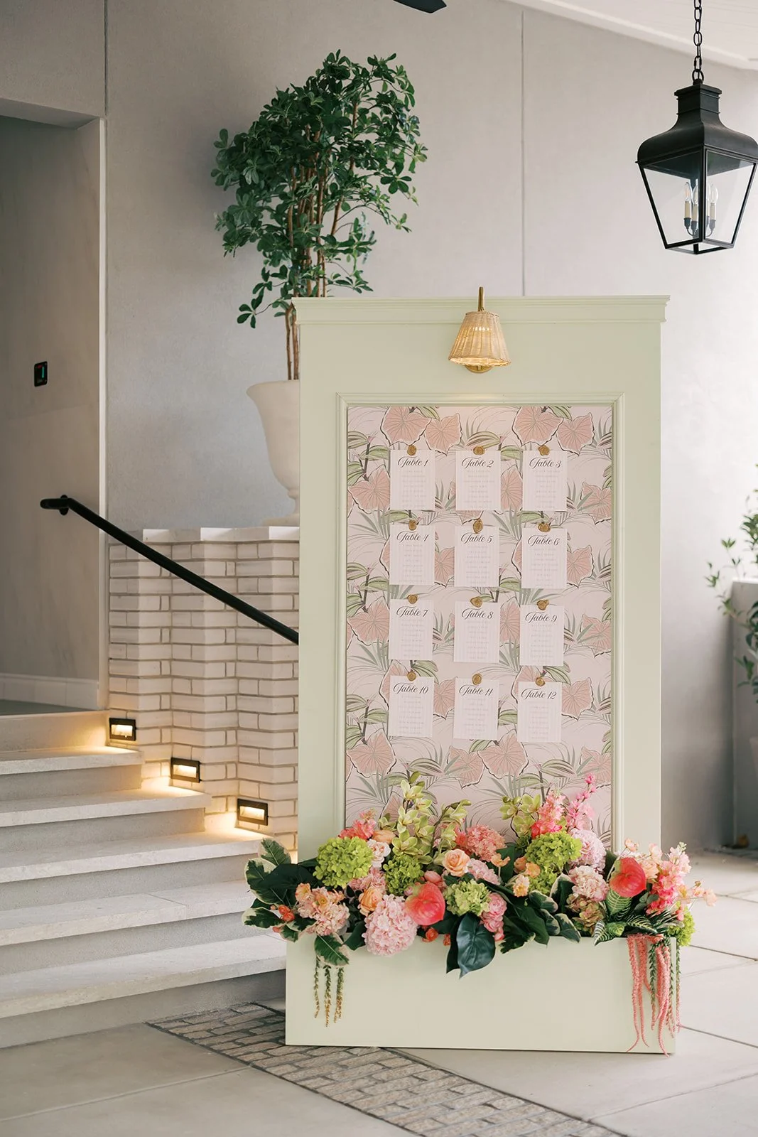 A large floral seating chart display for a wedding, decorated with pink and green flowers, placed at the bottom of a white frame with map-style seating cards, inside a decorated venue with stairs, potted plants, and a hanging light fixture.