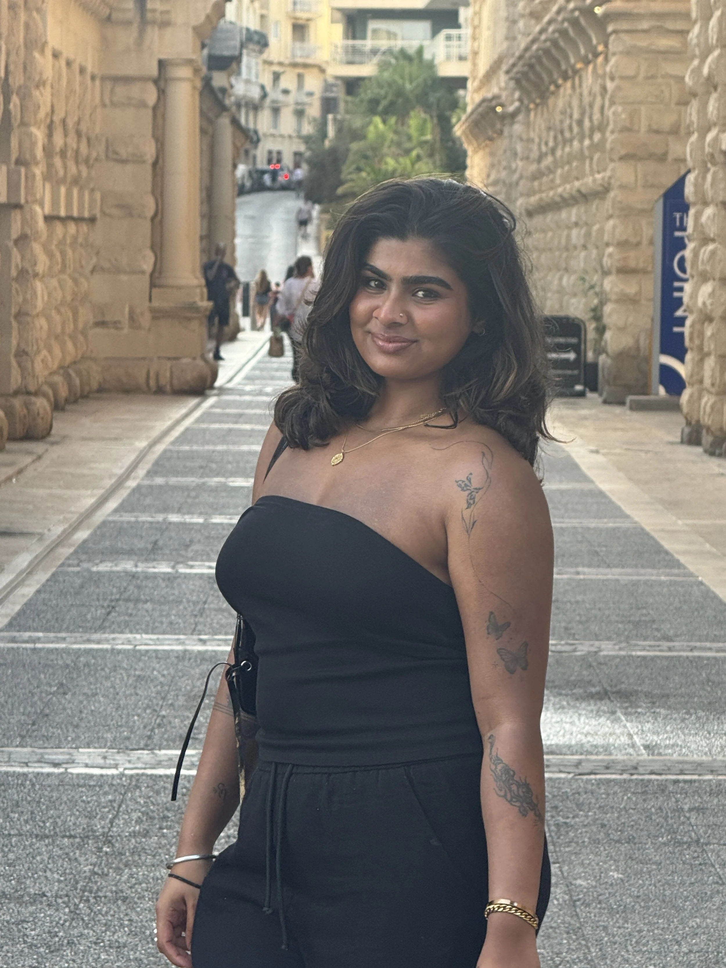 A woman with dark, wavy hair and tattoos on her arm, wearing a black off-shoulder top and gold jewelry, standing on a cobblestone street between stone buildings, with other people walking in the background.