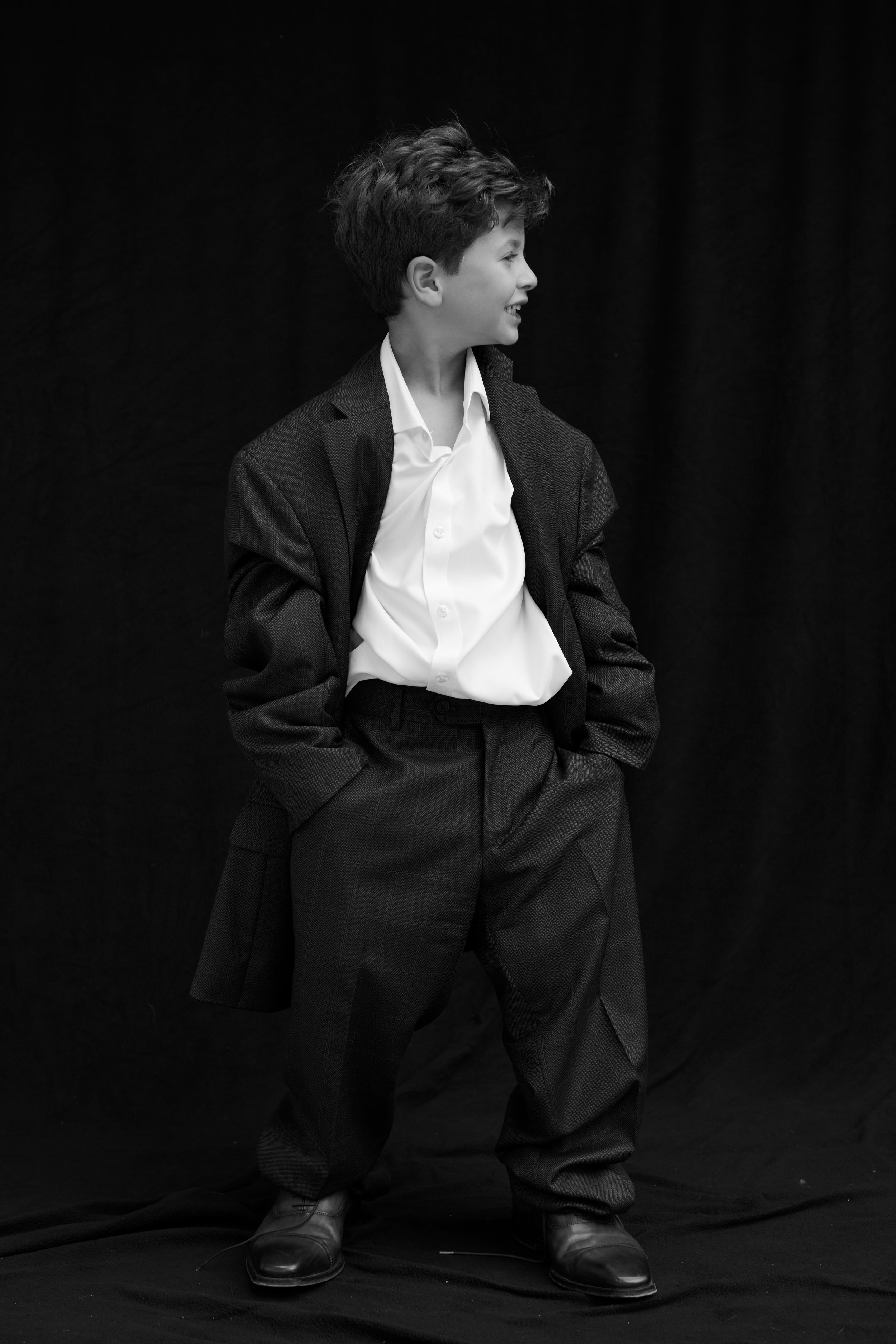 A young boy in a white shirt, dark checked trousers, and a dark blazer, with hands in pockets, standing against a black background, looking to the right and smiling.