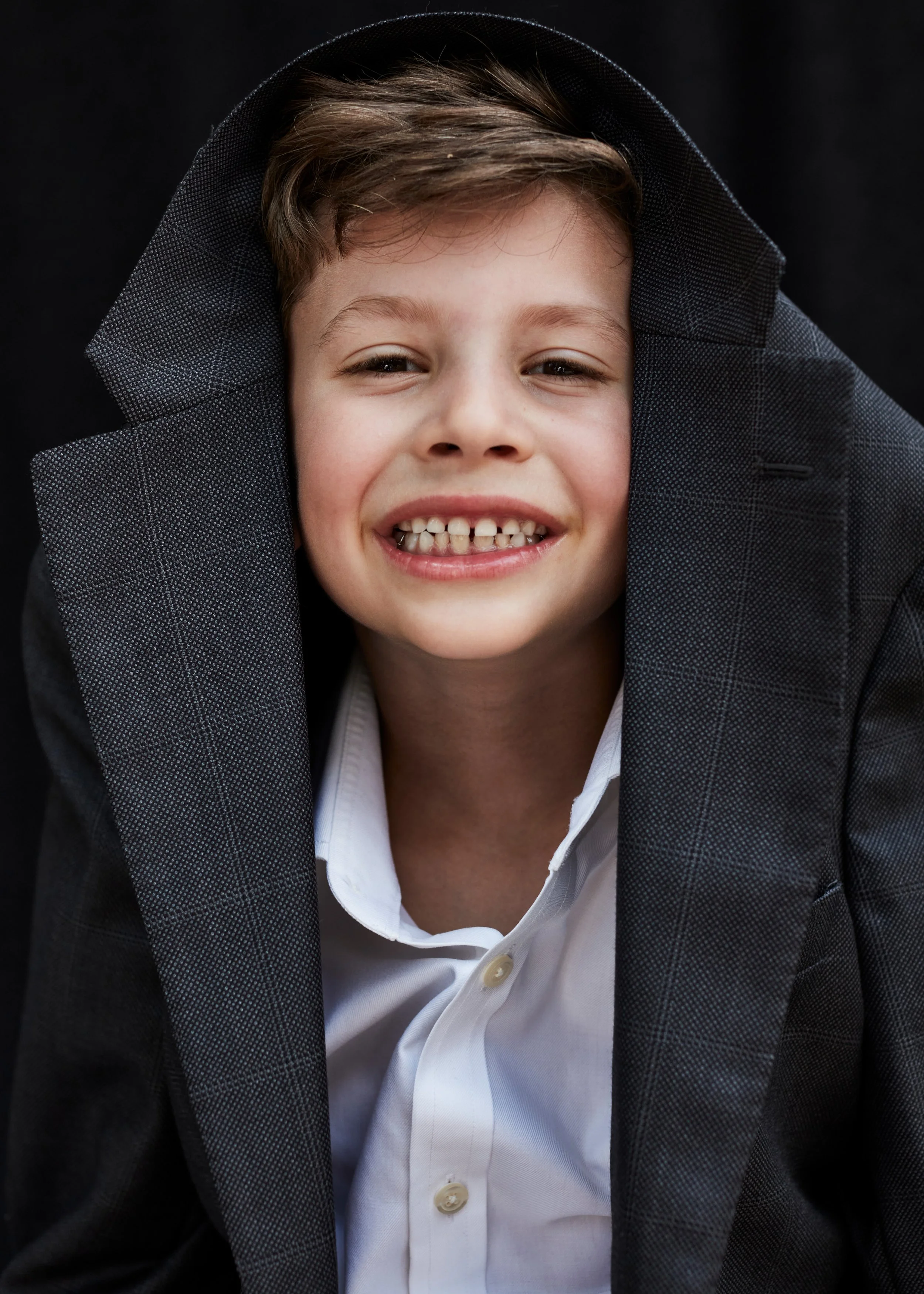 Close-up portrait of a smiling young boy with curly hair, wearing a dark blazer, white shirt, and a jacket with a checkered pattern, against a black background.