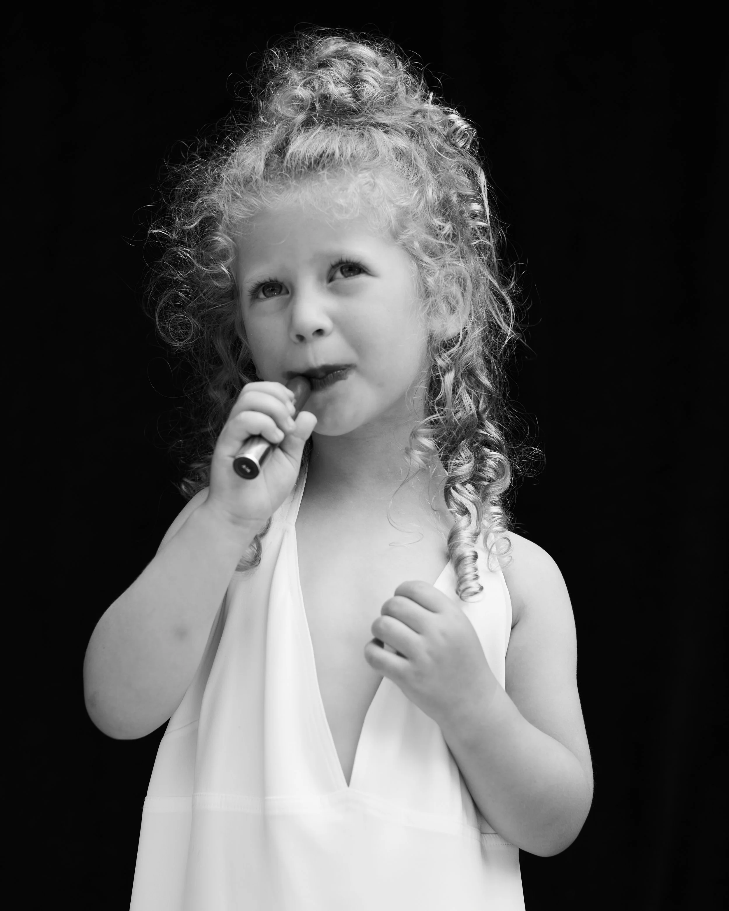 A young girl with curly hair, wearing a sleeveless top, holding a small object near her mouth against a black background.