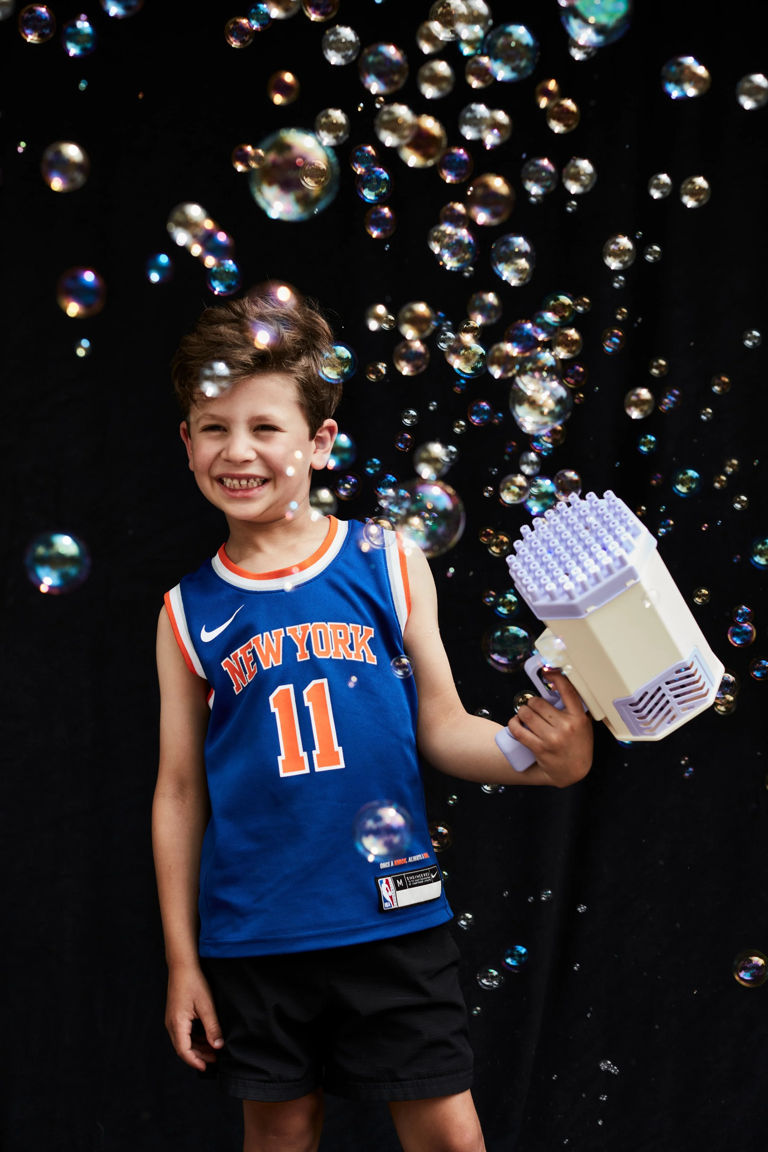 A young boy wearing a New York Knicks basketball jersey is standing in front of a black background, holding a bubble gun, with soap bubbles floating around him and a big smile on his face.