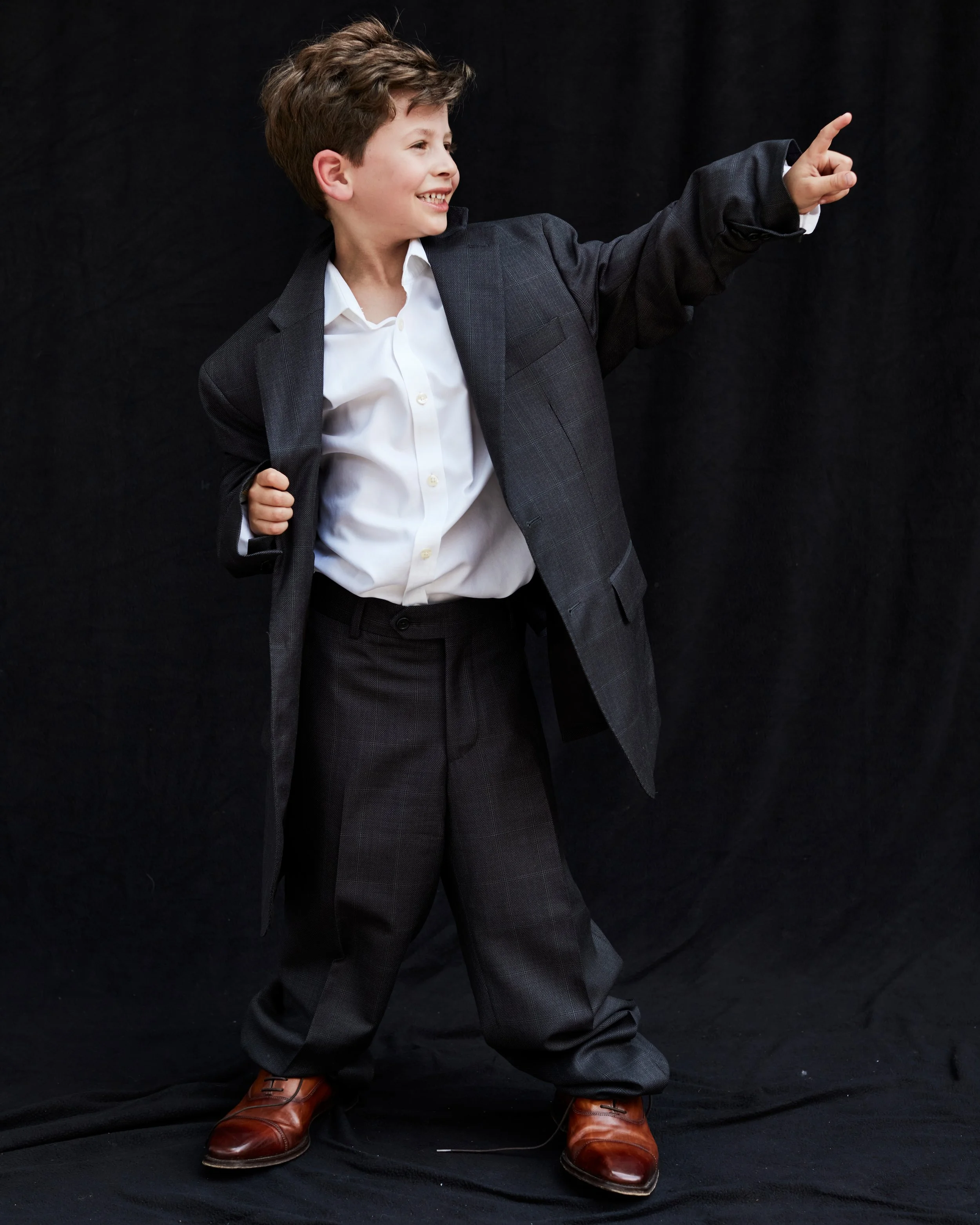 A young boy with brown hair dressed in a white shirt, a gray blazer, and matching gray pants, standing against a black background. He is smiling and pointing upward with his right hand while holding a black jacket over his left shoulder. He is wearing brown dress shoes.