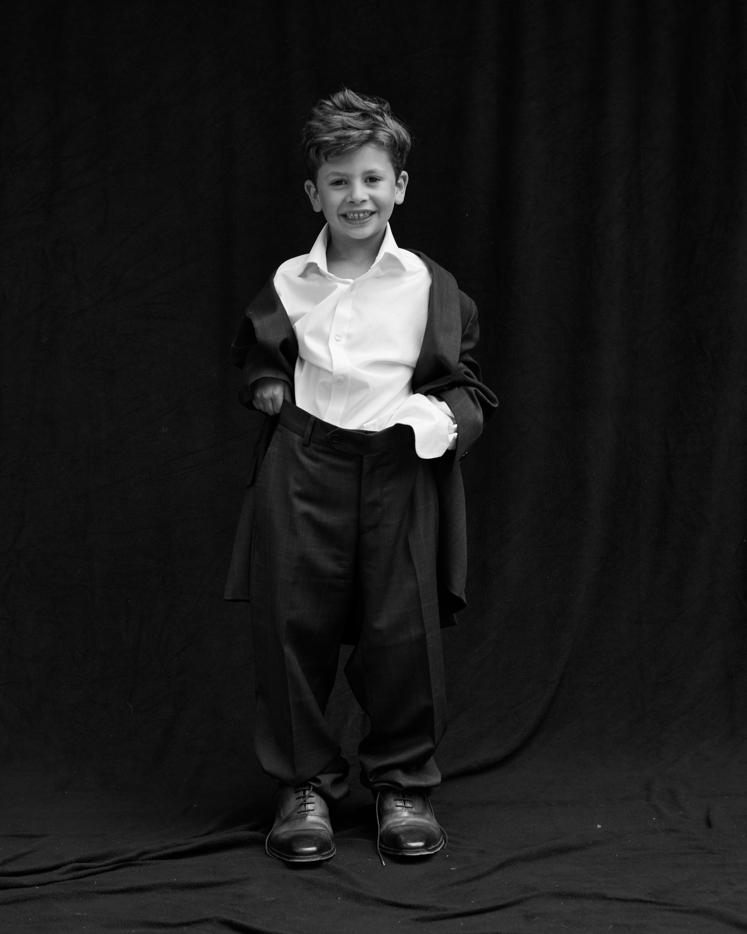 A young boy in dress clothes standing against a black background, smiling and looking at the camera.