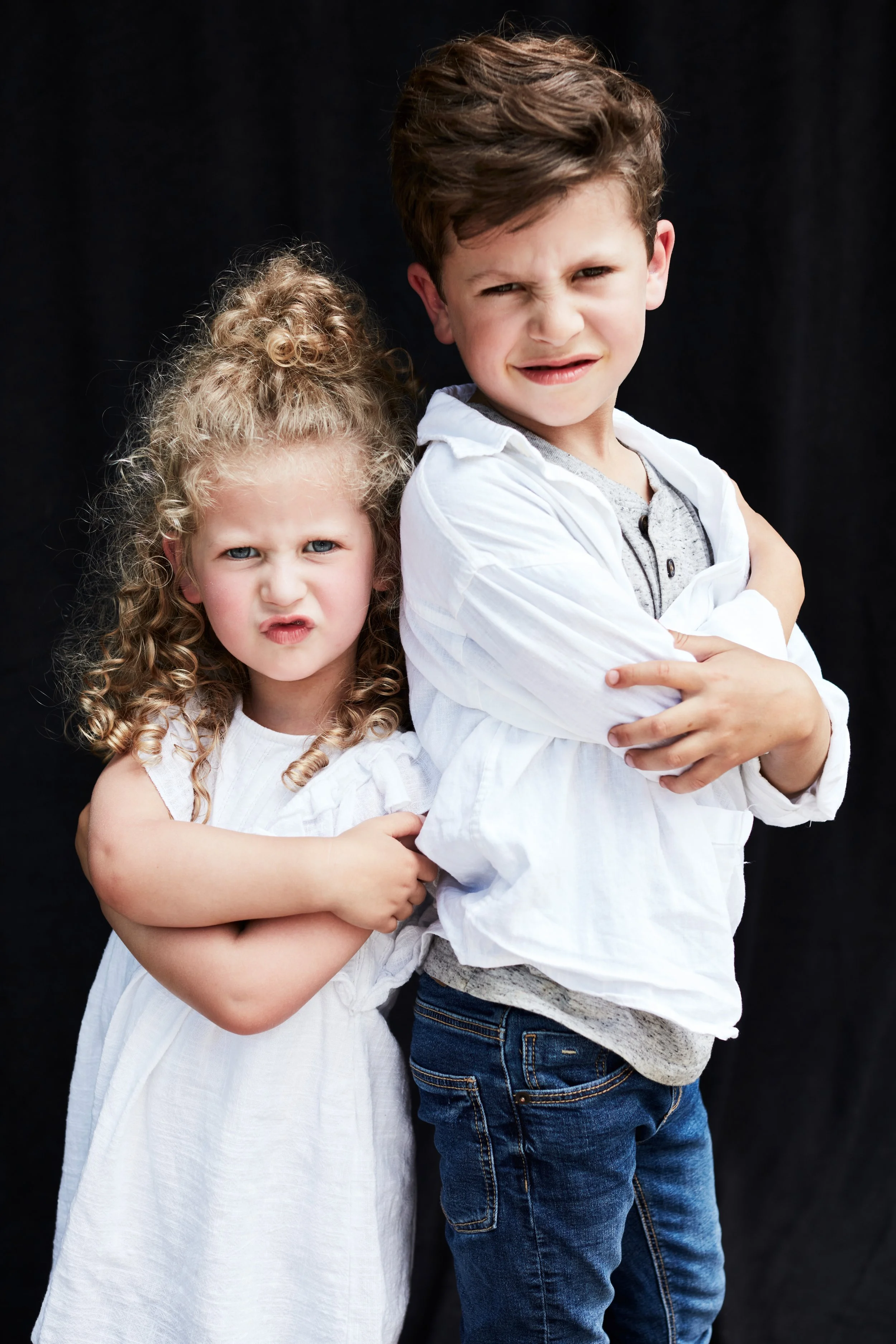 A boy with crossed arms and a girl hugging him from behind, both wearing white clothing, posing against a black background with playful facial expressions.