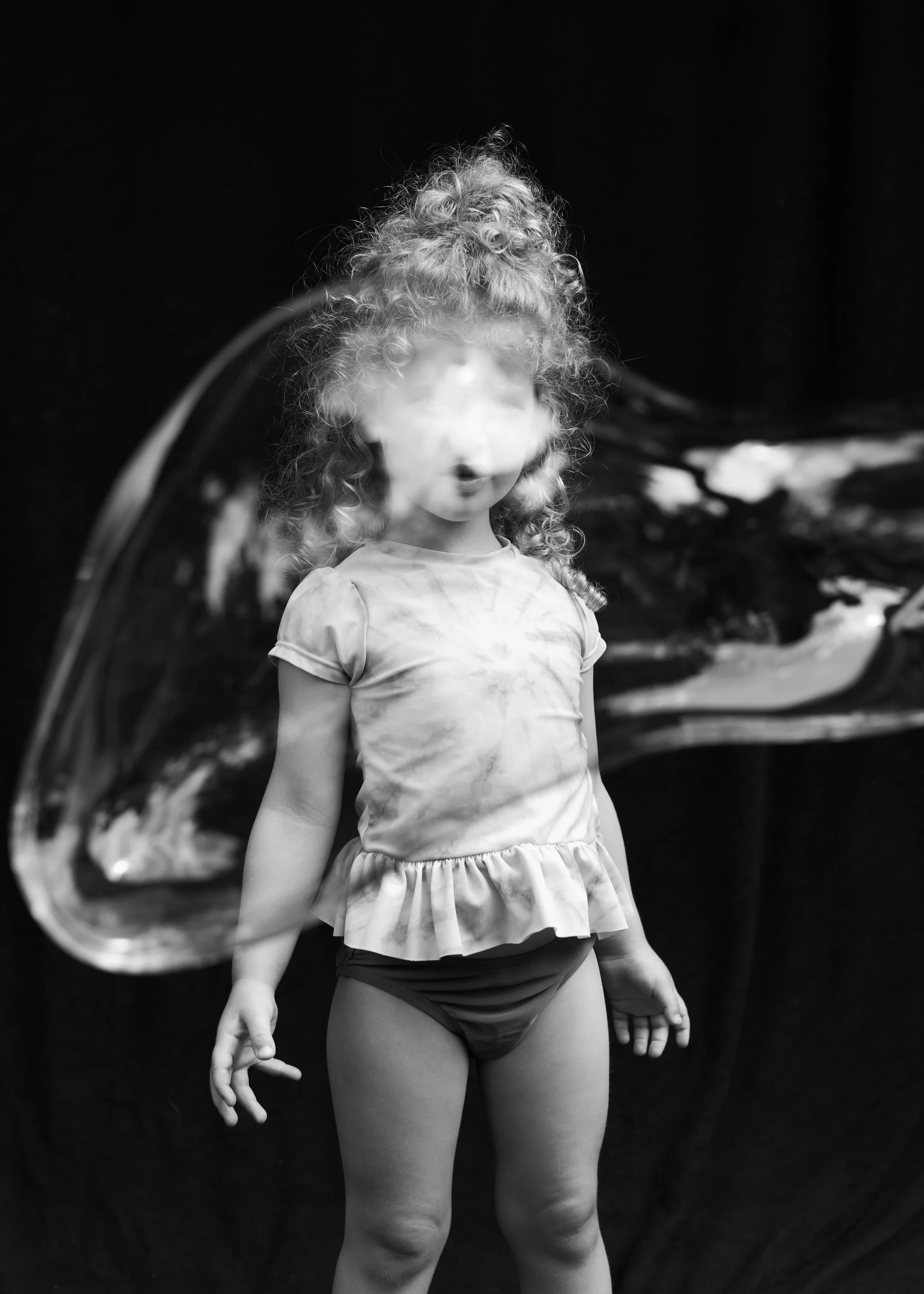 Black-and-white photo of a young girl with curly hair wearing a t-shirt with ruffled sleeves and shorts, standing outdoors in front of a reflective surface, possibly a mirror, with a dark background.