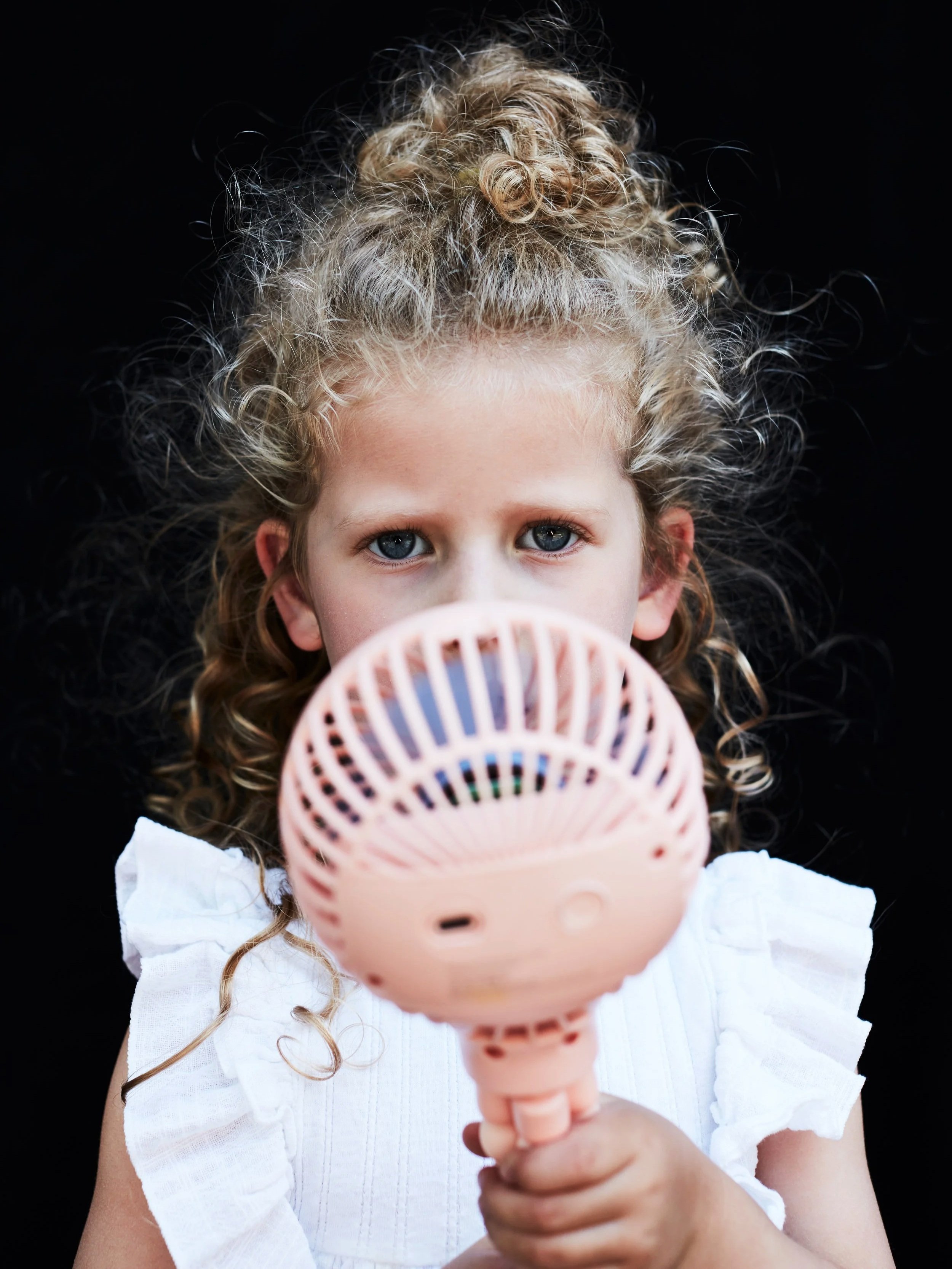 A young girl with curly blonde hair and blue eyes holding a pink cricket microphone in front of her face.