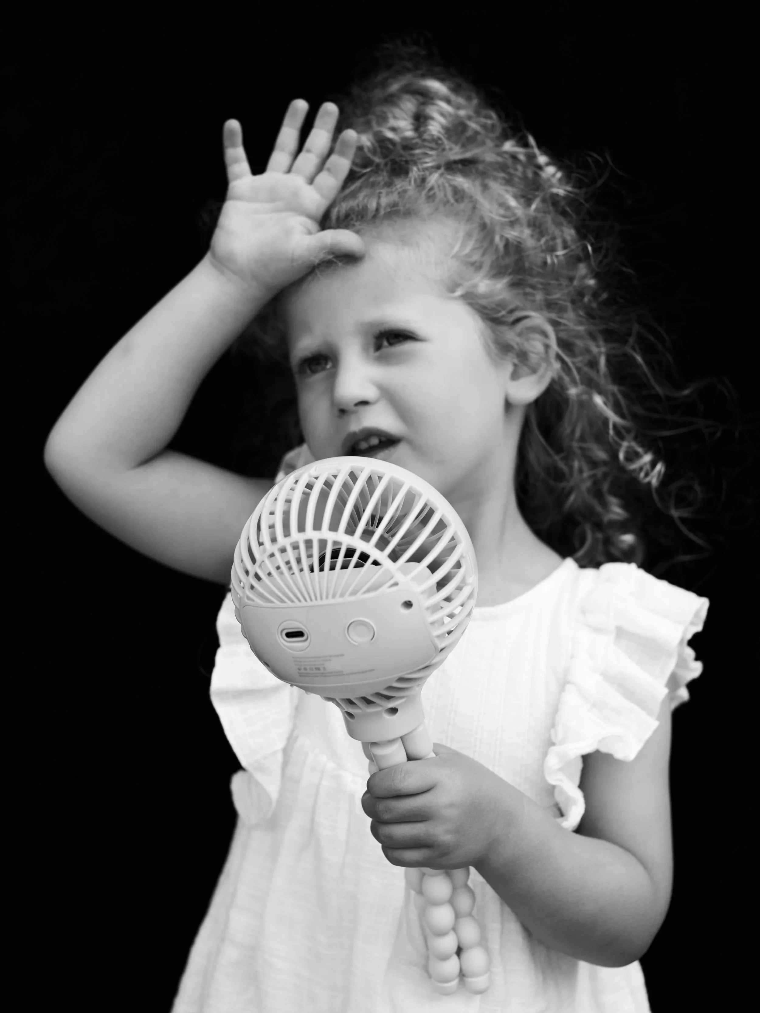 A young girl with curly hair holding a handheld fan and a small portable fan, with her hand raised near her face, against a black background.
