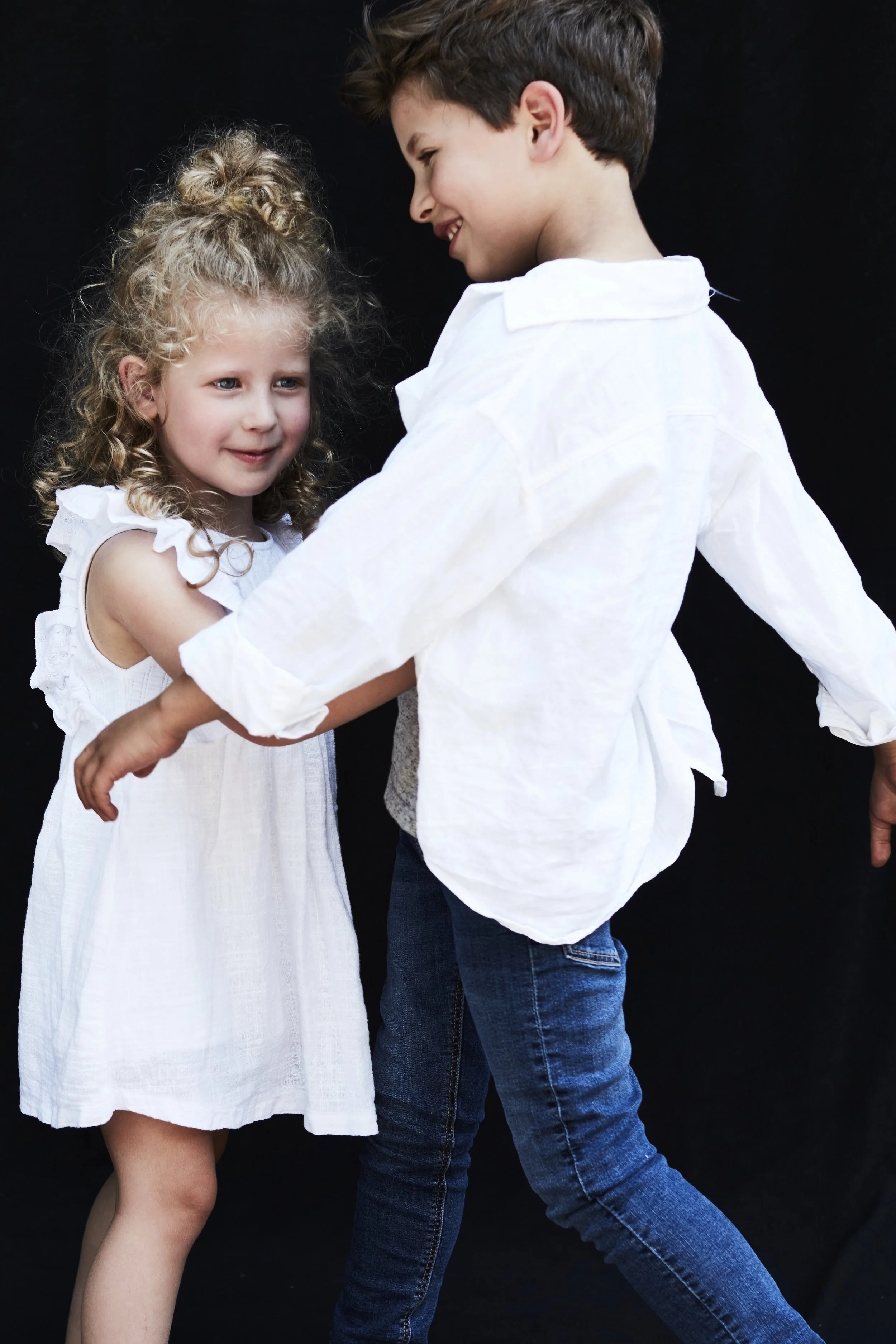 A young boy and girl are dancing and smiling together against a black background. The girl has curly blonde hair and is wearing a white dress, while the boy has brown hair and is wearing a white shirt and blue jeans.