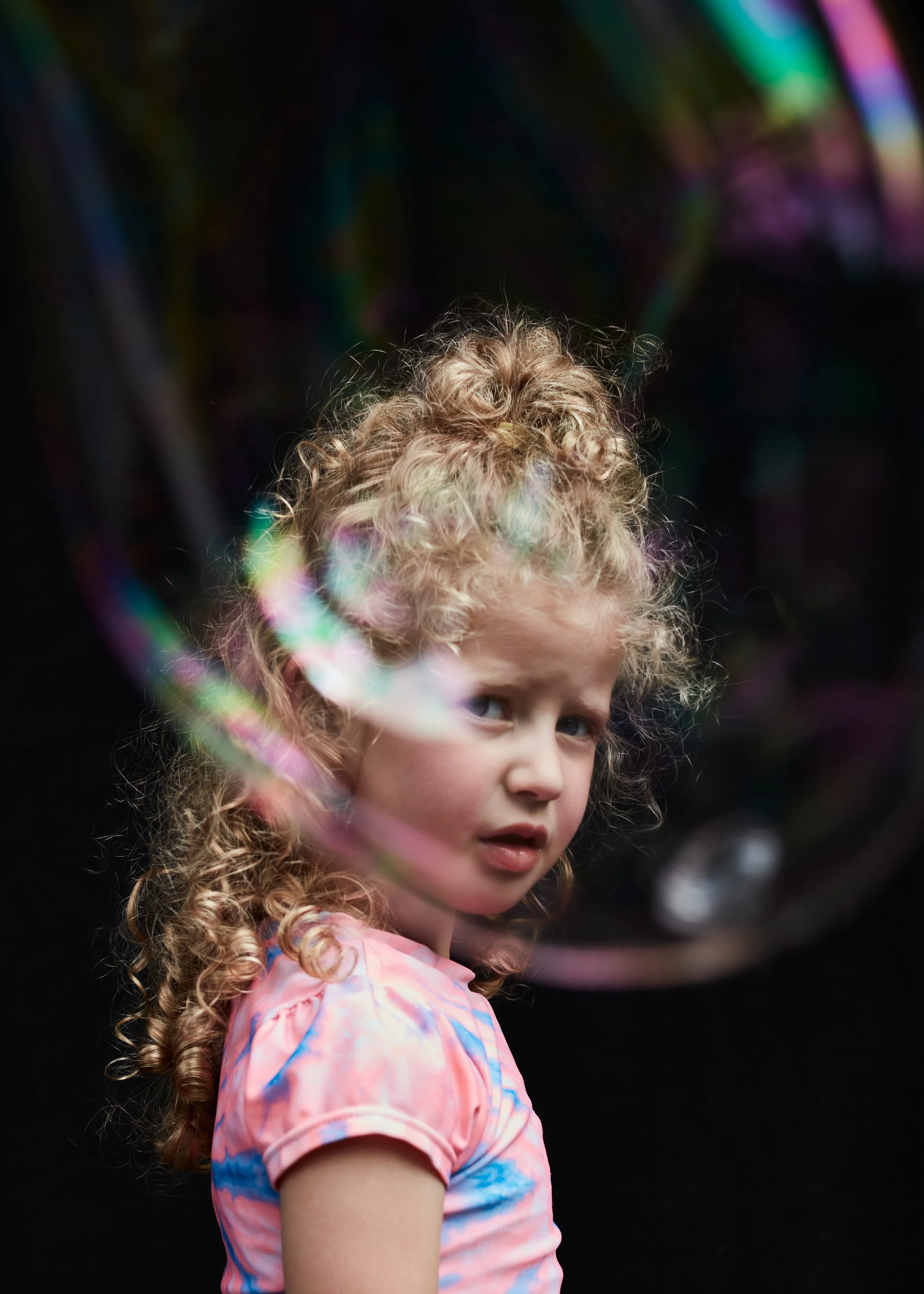 A young girl with curly blonde hair and blue eyes looking over her shoulder, wearing a colorful tie-dye shirt, with a dark background and rainbow-colored light reflections.