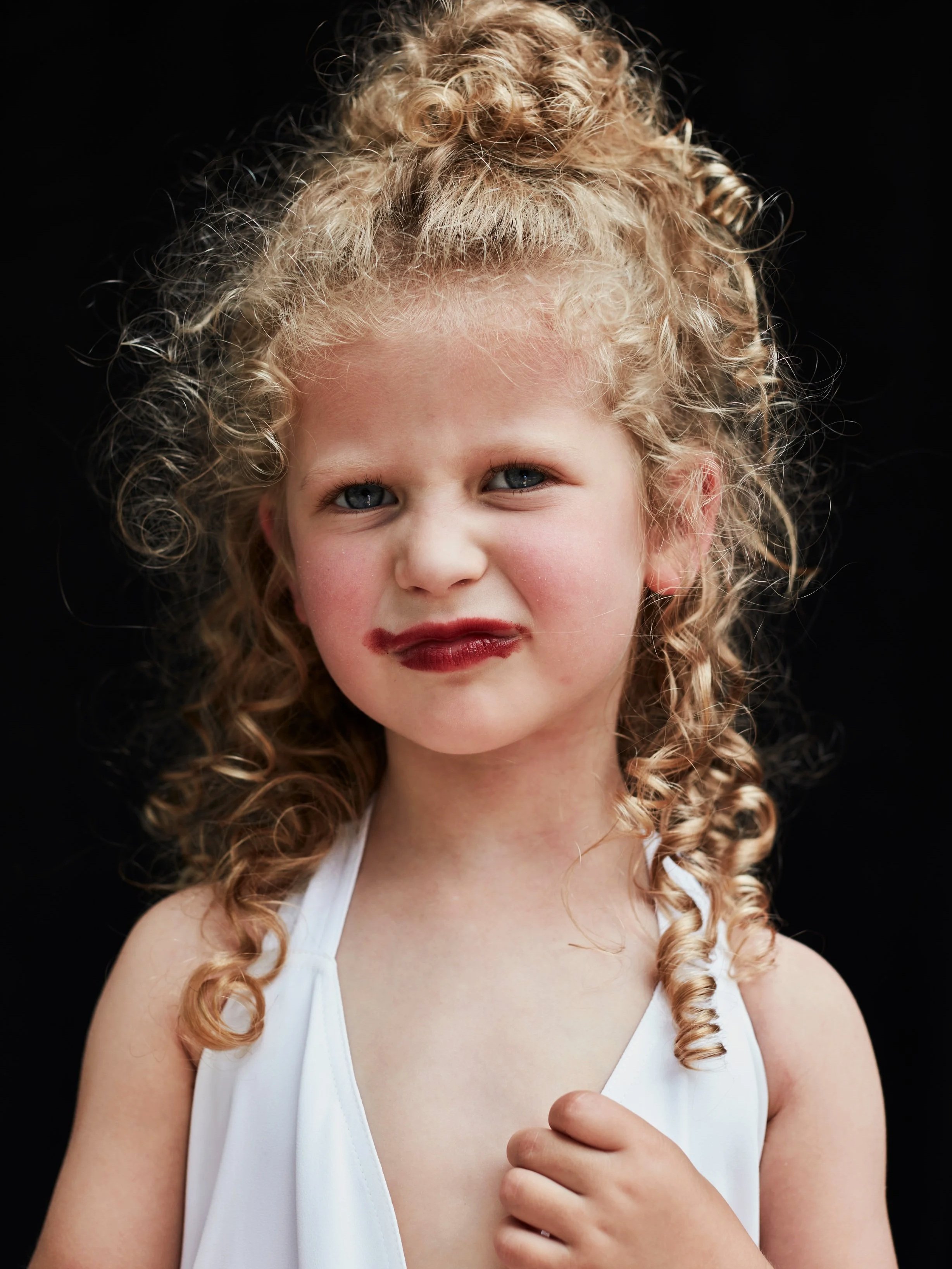 A young girl with curly blonde hair styled in a high bun and loose curls down her shoulders, wearing a white sleeveless top, against a plain black background.