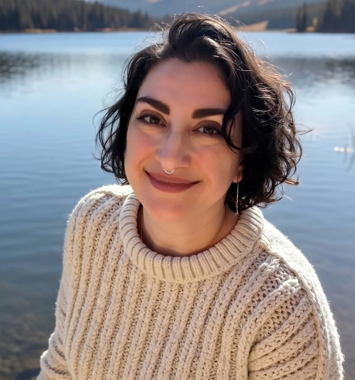 A woman with short, wavy dark hair and a septum piercing smiles at the camera while standing near a body of water with a forested shoreline and hills in the background, wearing a chunky cream-colored knit sweater.
