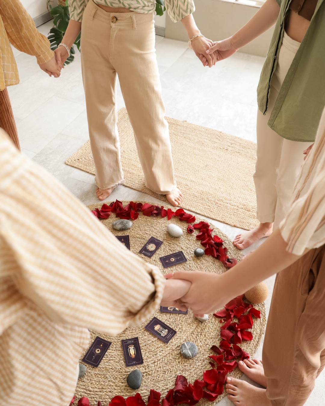 A group of people holding hands in a circle for a Tarot card reading or spiritual ritual, with a spread of Tarot cards, rocks, and red rose petals on a woven rug.