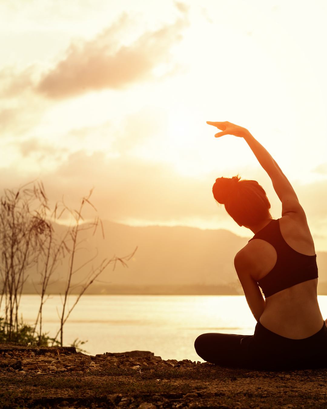 A woman practicing yoga outdoors at sunset, sitting cross-legged by water with mountains in the background, doing a side stretch with her arm overhead.