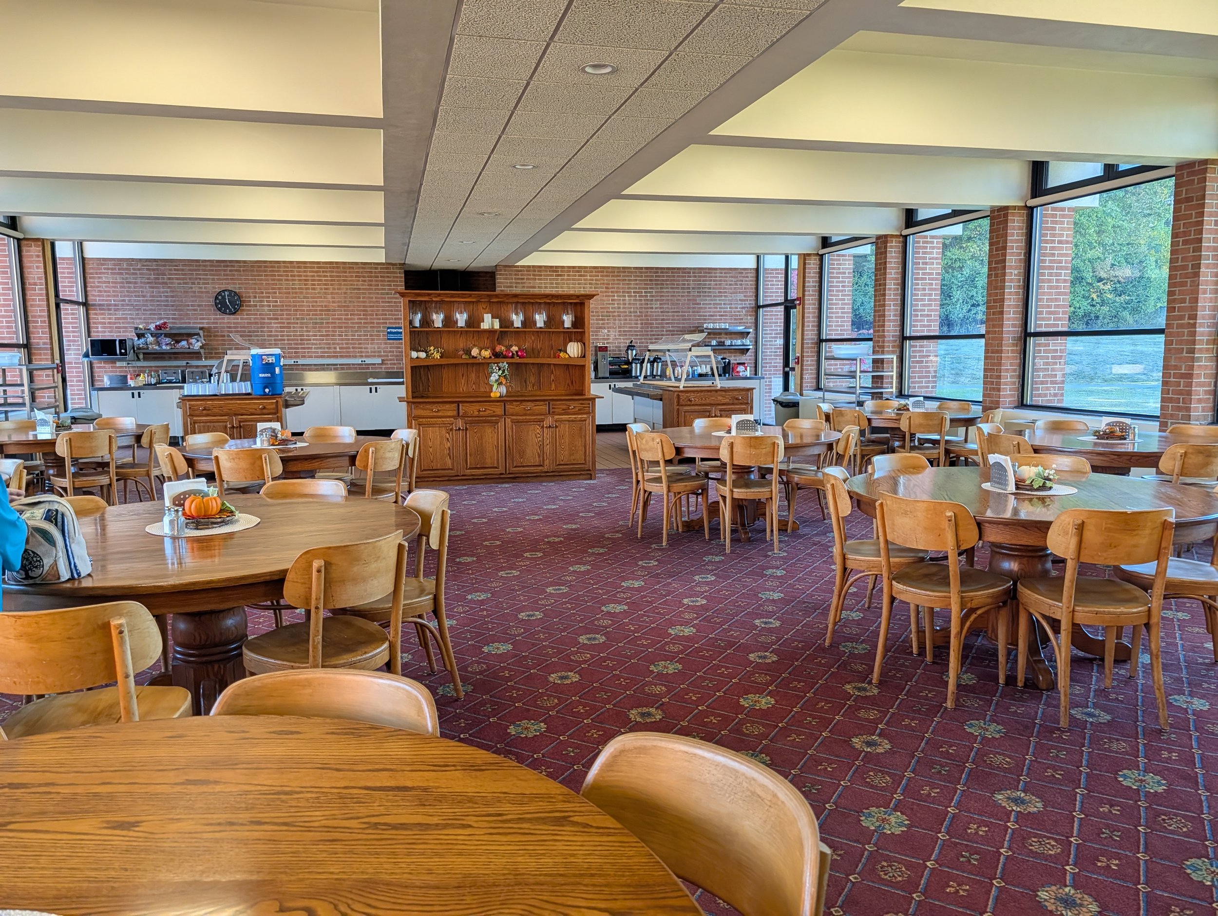 Interior of a cafeteria or dining area with wooden tables and chairs, large windows, a brick wall, and a buffet or serving counter.